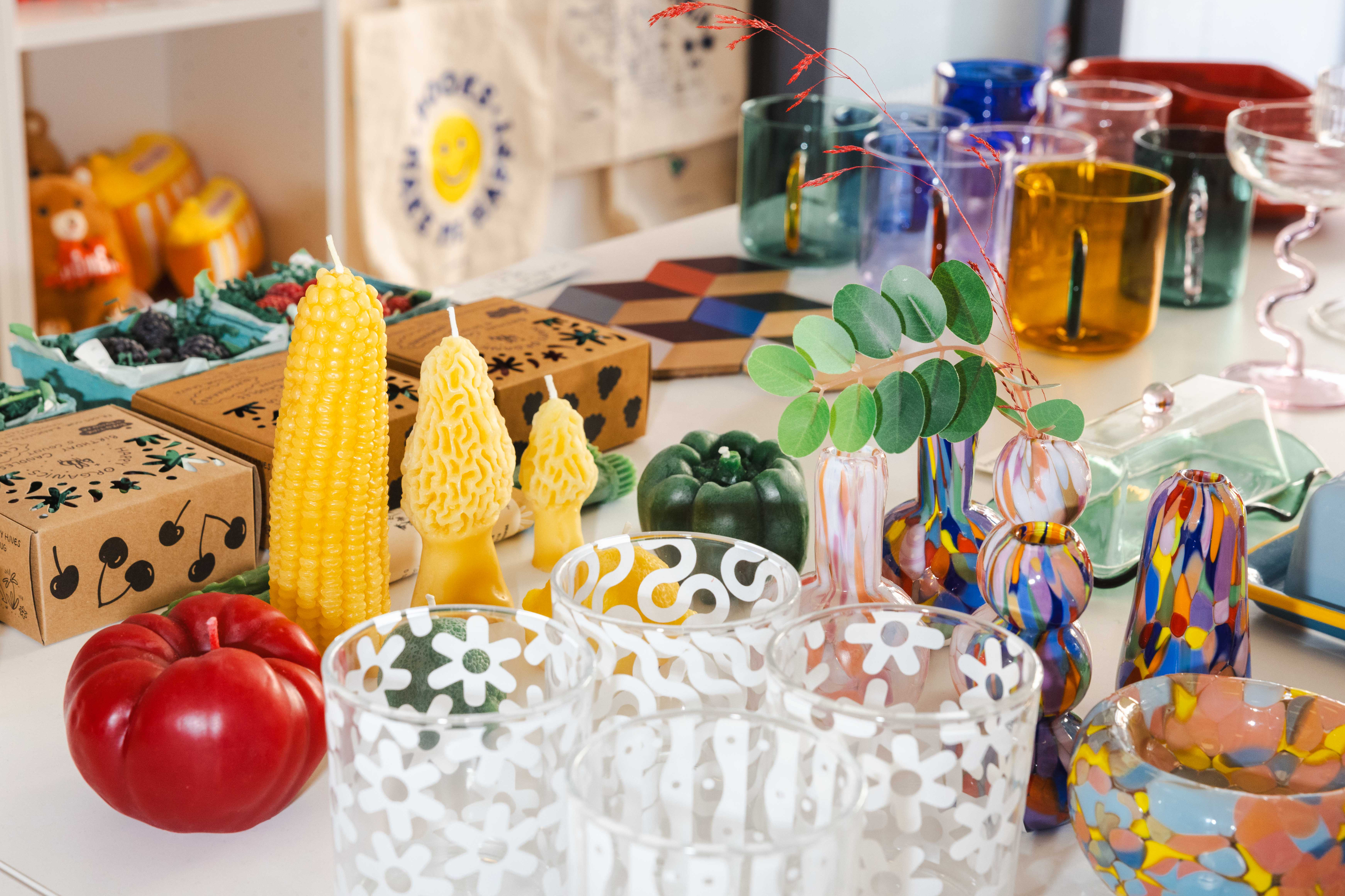 The image shows a variety of colorful glassware, decorative candles shaped like corn and trees, a red apple, and boxes with designs on a table.