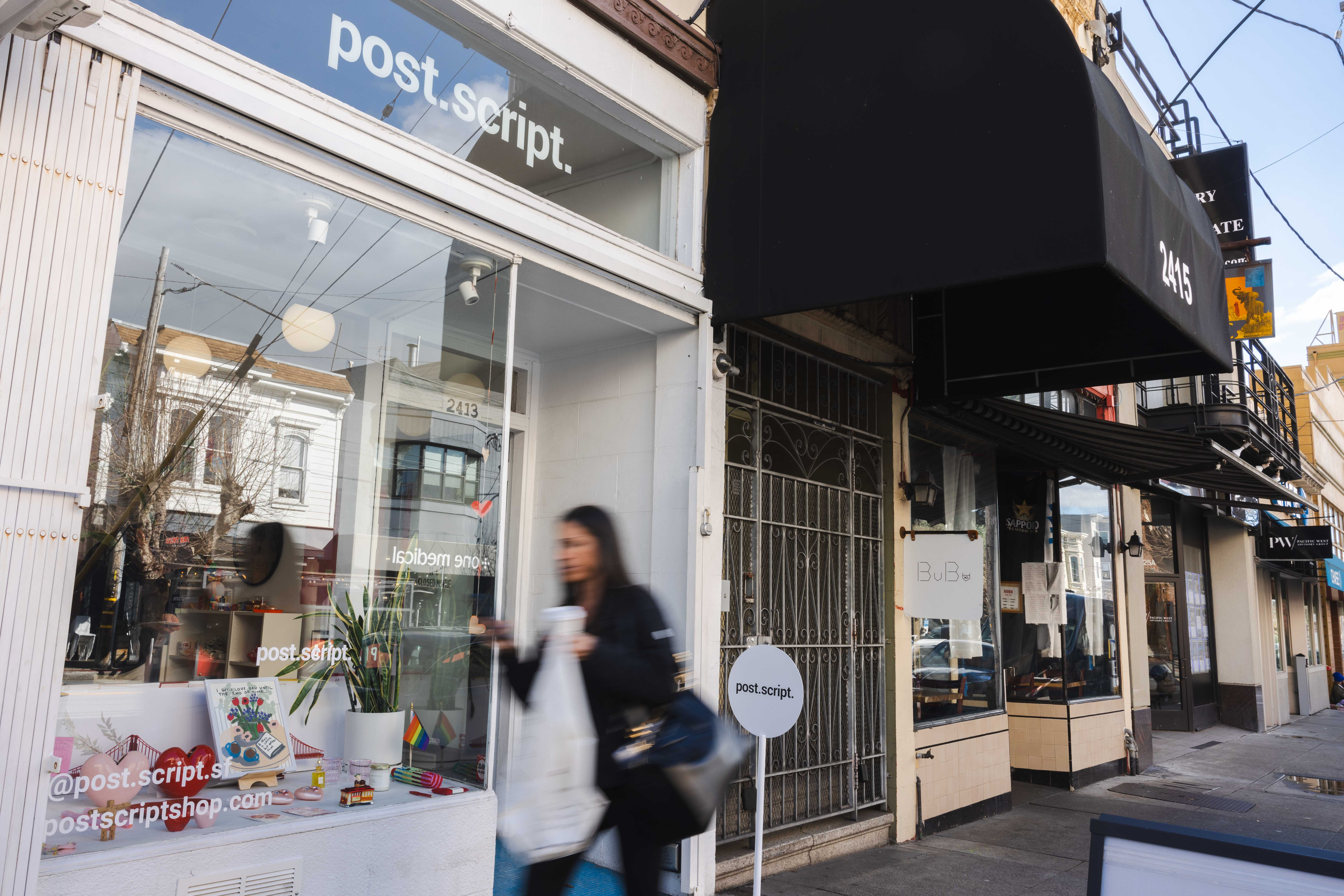 A storefront with the sign &quot;post.script&quot; above the entrance. A blurred person walks by holding a bag and drink. The window displays colorful items.