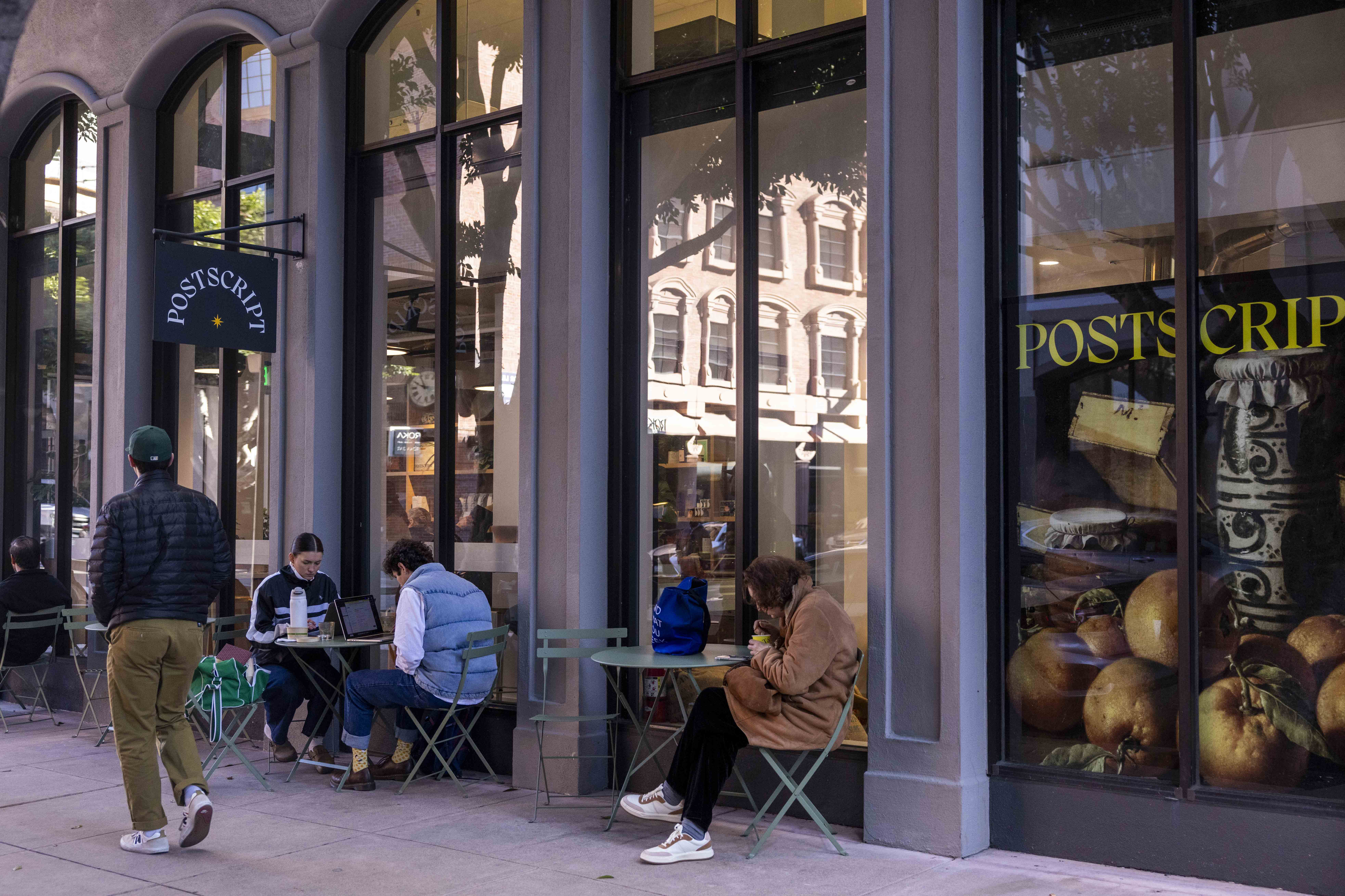 People are seated at small green tables outside a building with large windows. A sign reads &quot;Postscript.&quot; A person walks by, and a window displays fruit images.