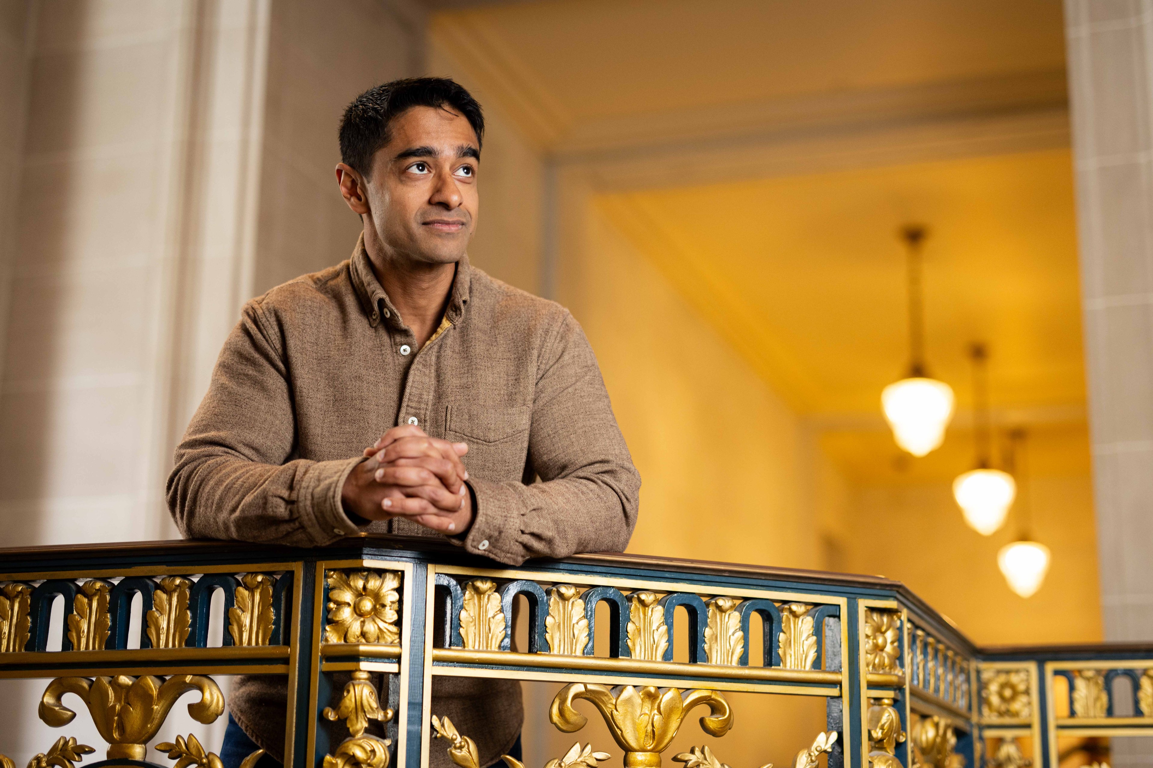A person in a brown shirt leans on an ornate, gold-accented railing indoors. Three ceiling lights hang in the background, illuminating the space warmly.