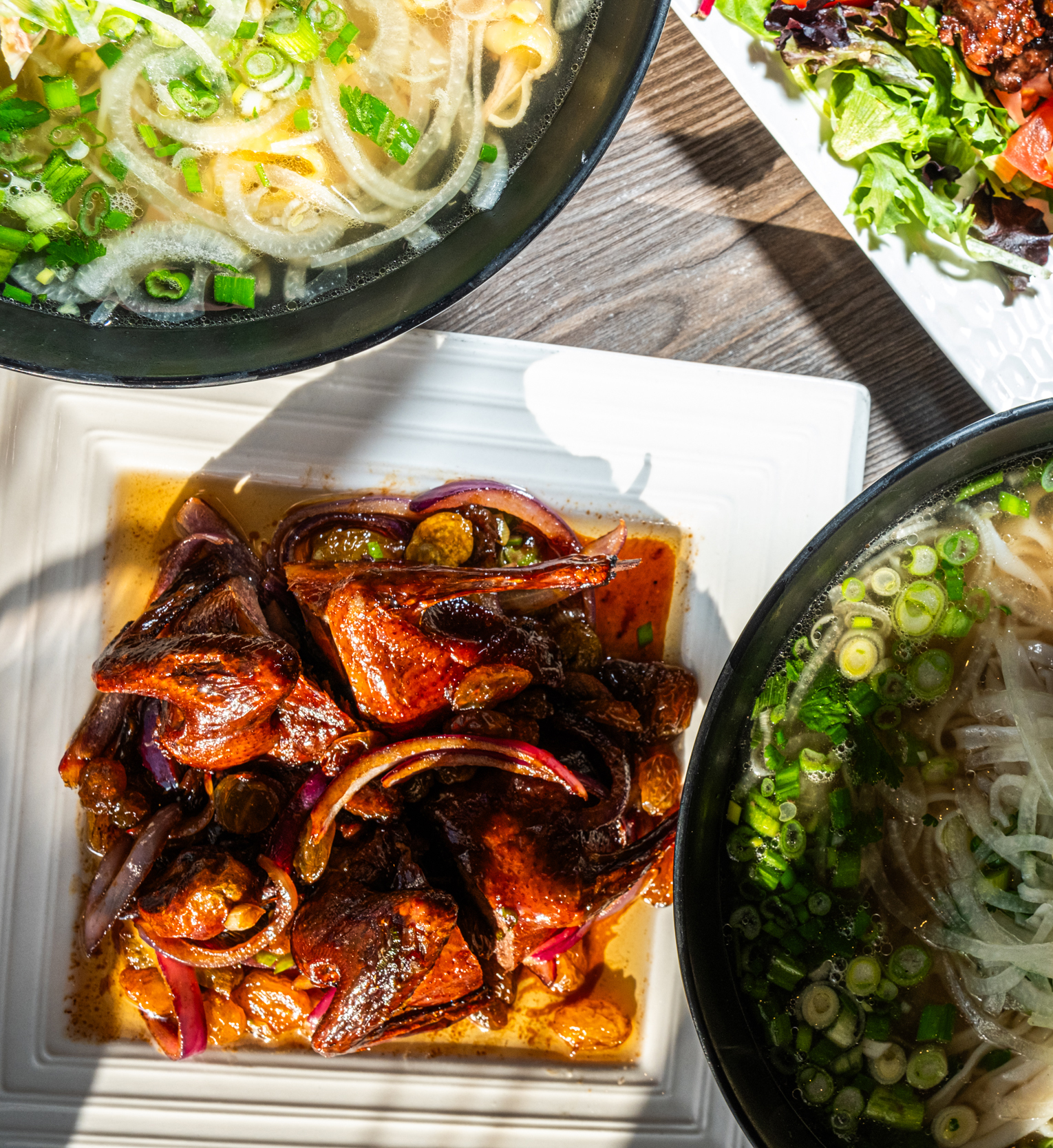 Two bowls of noodle soup with green onions and a dish of glazed meat with onions are on a table, alongside a plate of mixed salad greens.