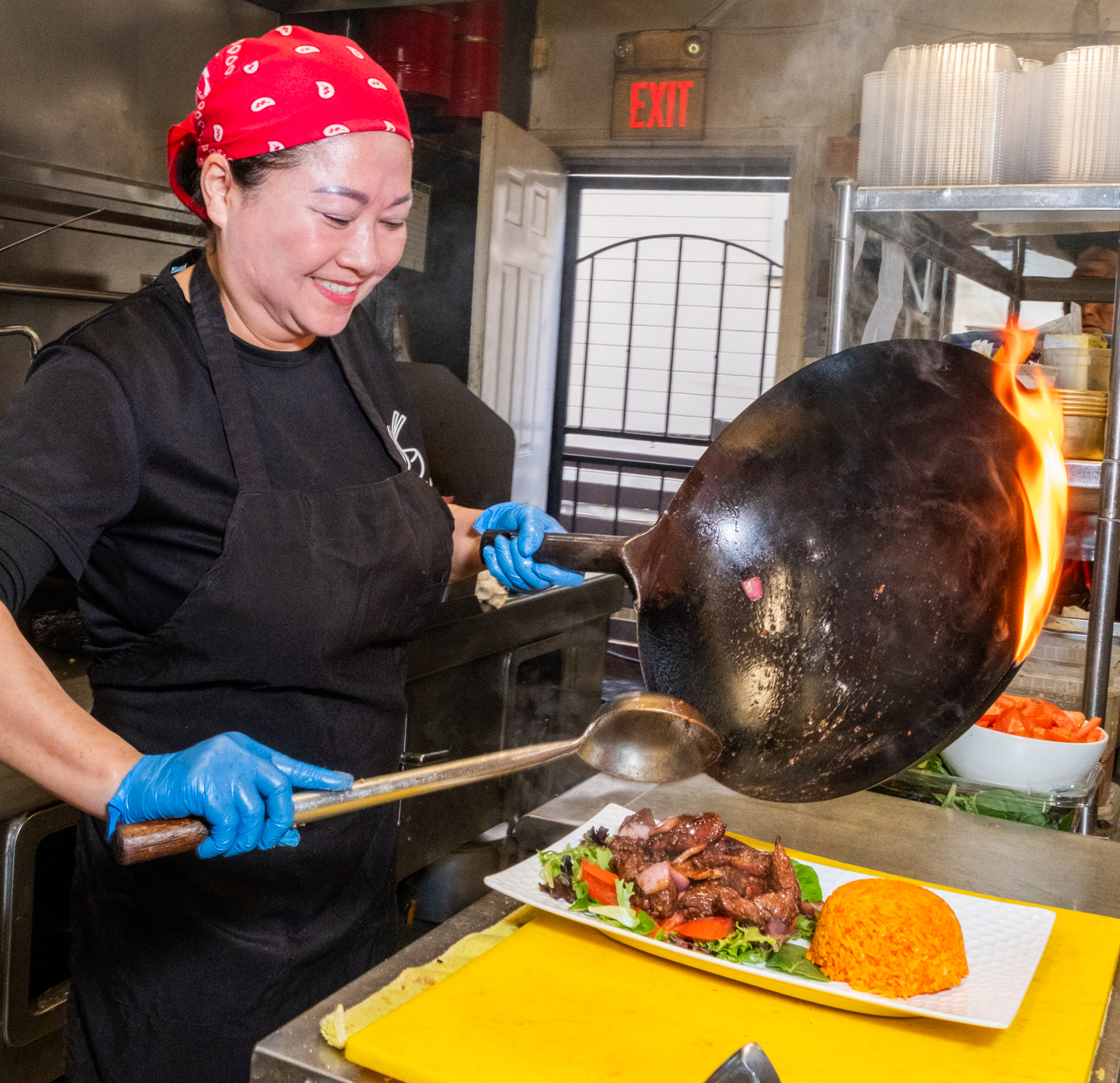 A chef wearing a red bandana and blue gloves smiles while cooking. She holds a flaming wok above a plate of food with rice on a yellow cutting board.