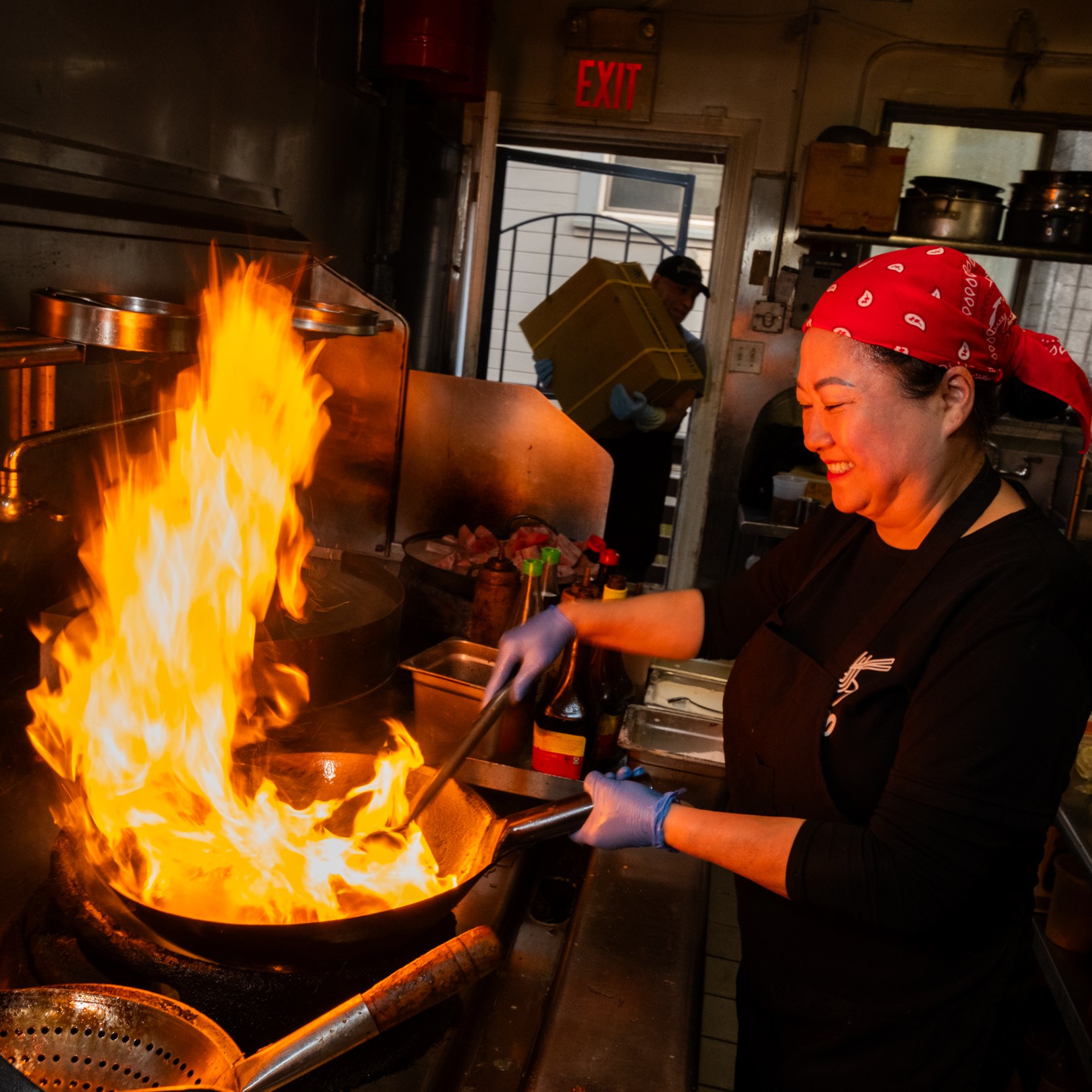A person in a red bandana is cooking in a kitchen, smiling while stirring food in a flaming pan. The kitchen has shelves with ingredients and an "EXIT" sign visible.