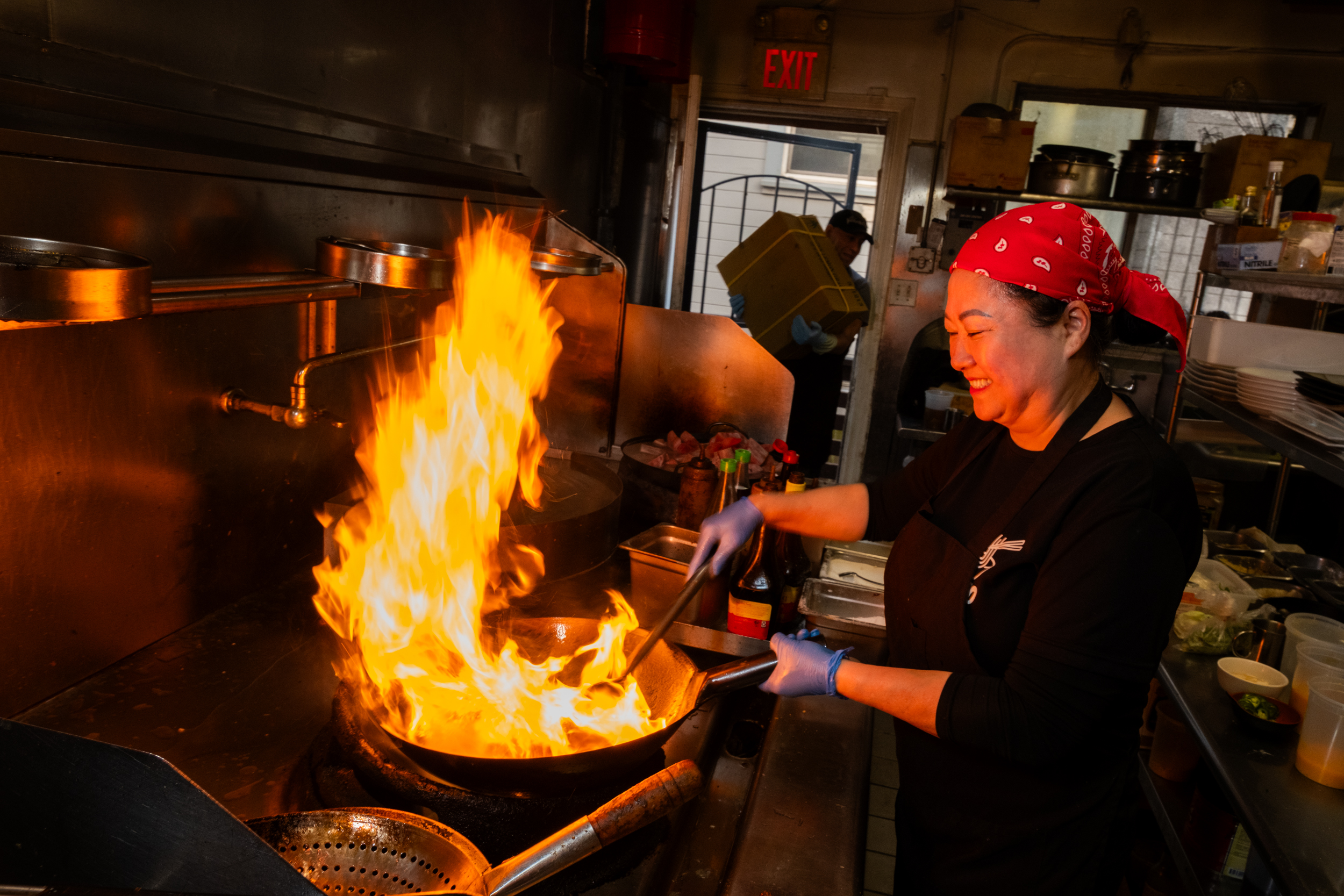 A person in a red bandana is cooking in a kitchen, smiling while stirring food in a flaming pan. The kitchen has shelves with ingredients and an "EXIT" sign visible.