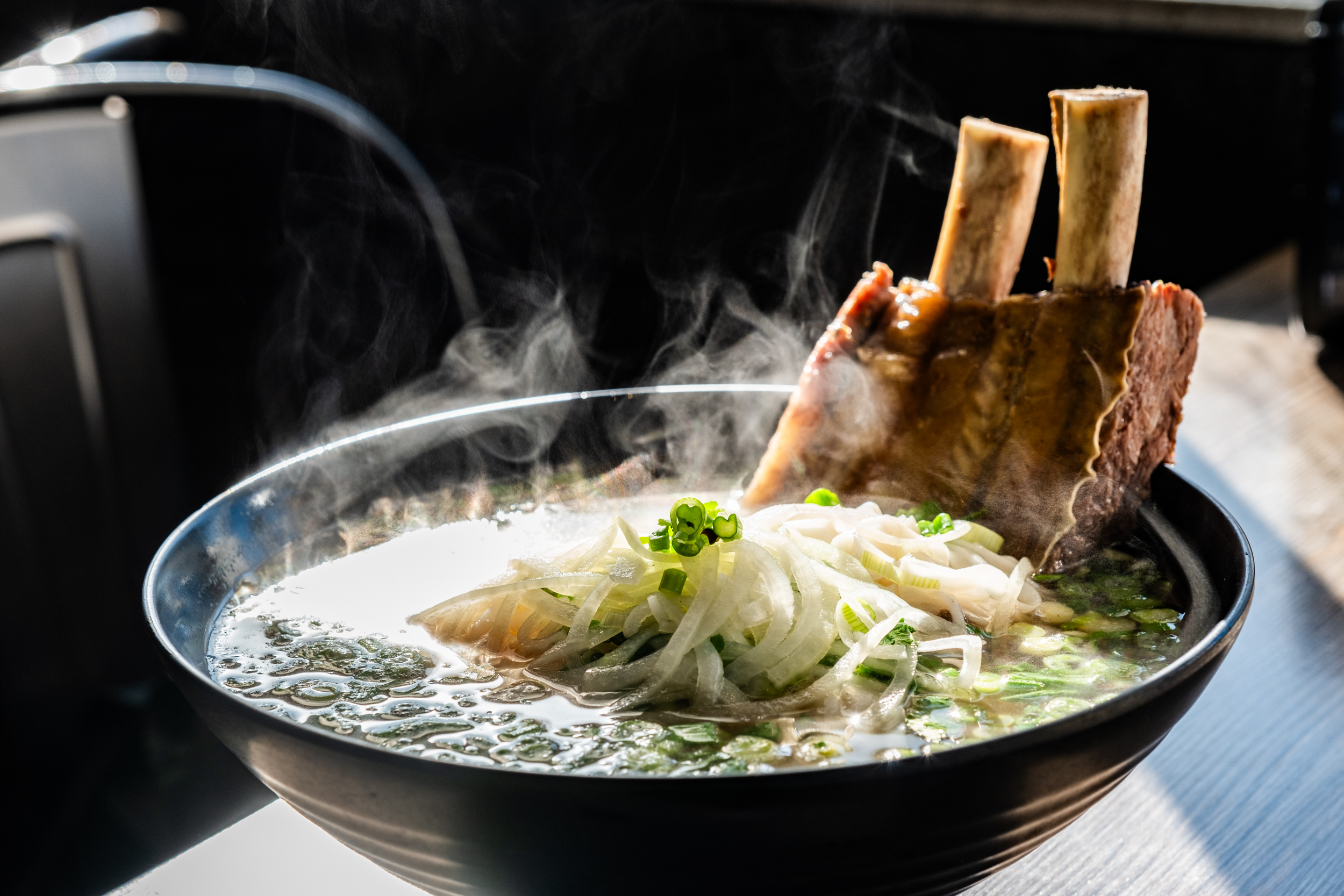 A steaming bowl of soup with a large beef bone, garnished with sliced onions and green onions, sits in a black bowl, with steam rising visibly.