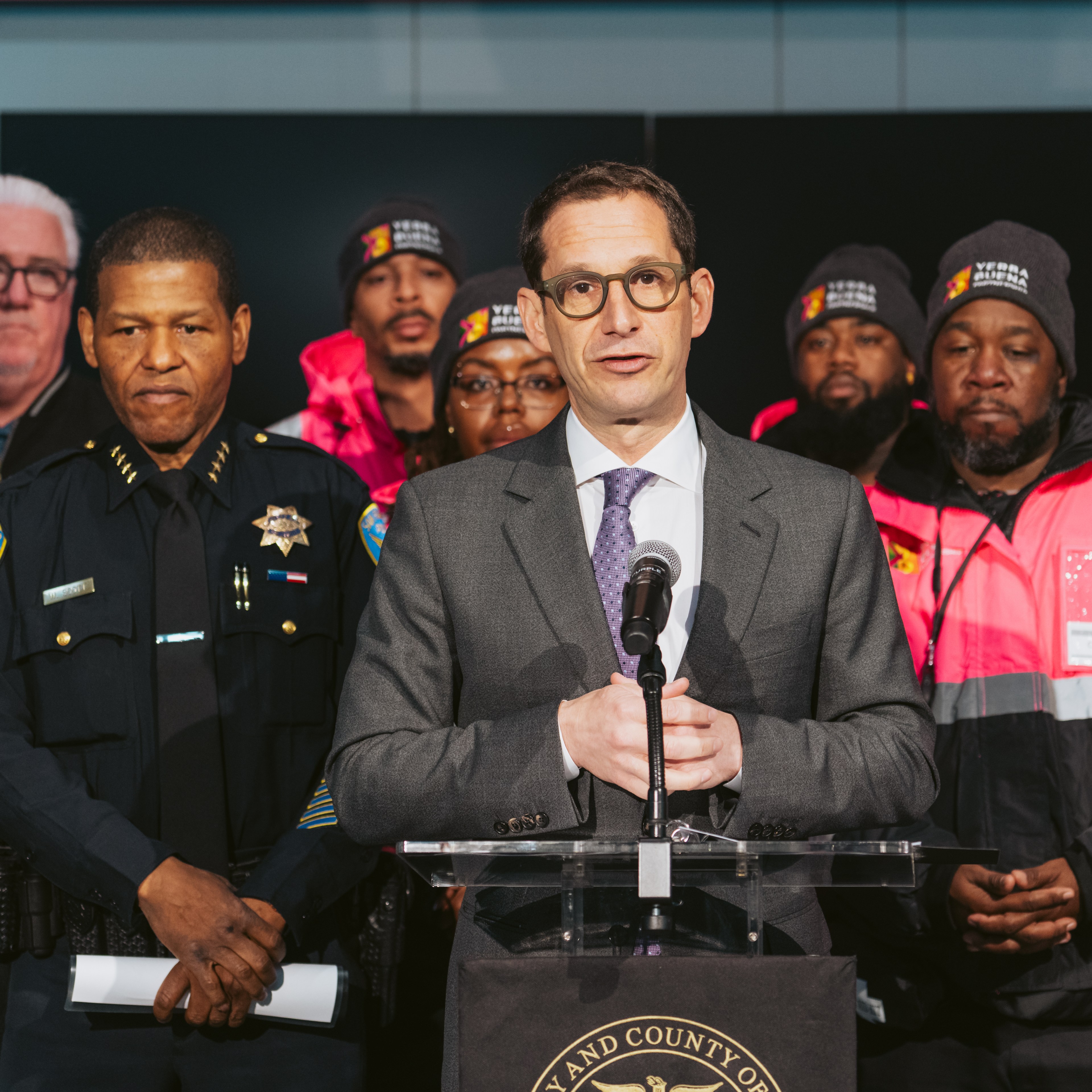 A man in a suit speaks at a podium, surrounded by uniformed officers and individuals in bright pink jackets, likely indicating a formal announcement or briefing.