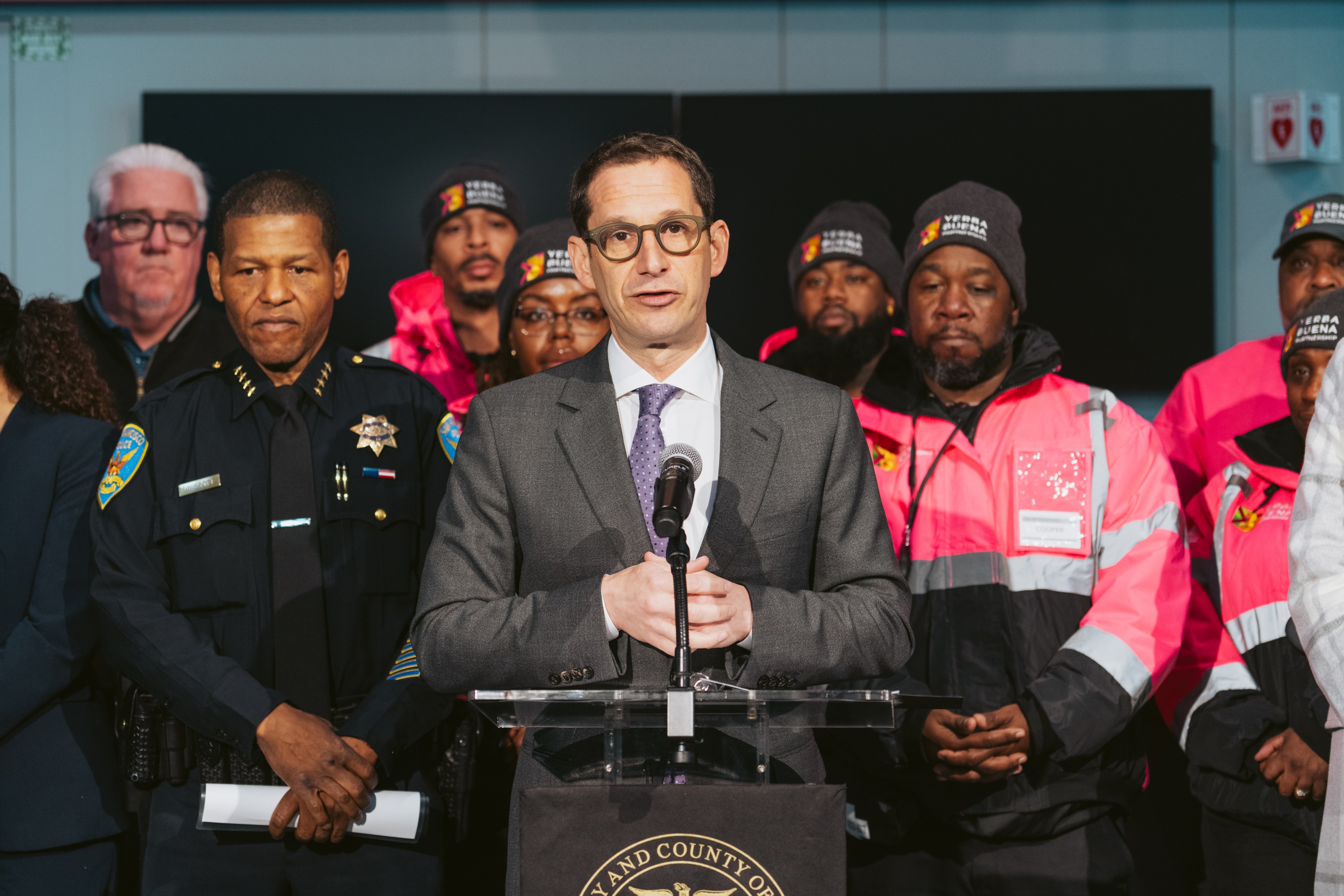 A man in a suit speaks at a podium, surrounded by uniformed officers and individuals in bright pink jackets, likely indicating a formal announcement or briefing.