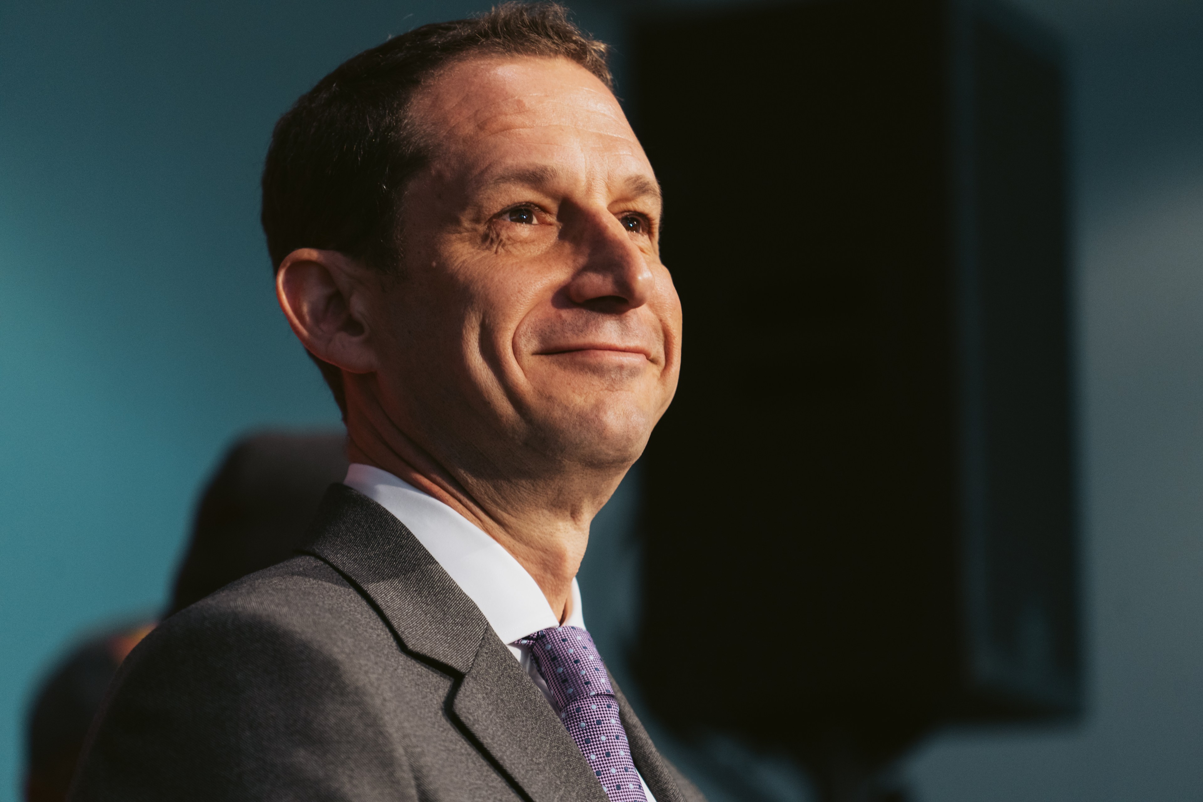 A man in a gray suit and purple tie smiles slightly, looking to the side. The background is a soft blue, with a dark, blurred shape behind him.