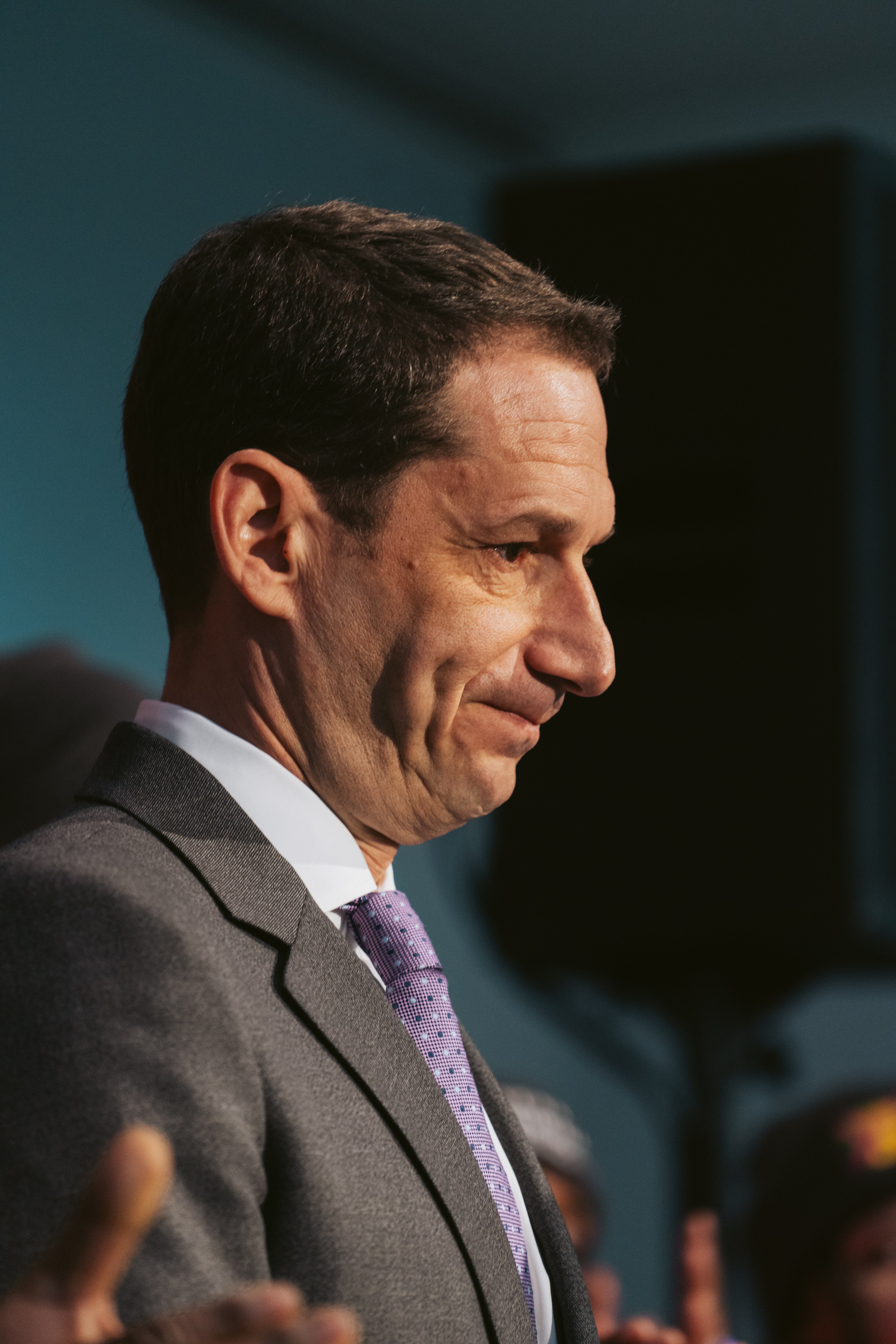 The image shows a man in a gray suit and purple tie, looking to the side with a slight smile. The background is a muted blue, and there's a shadowy hand visible.