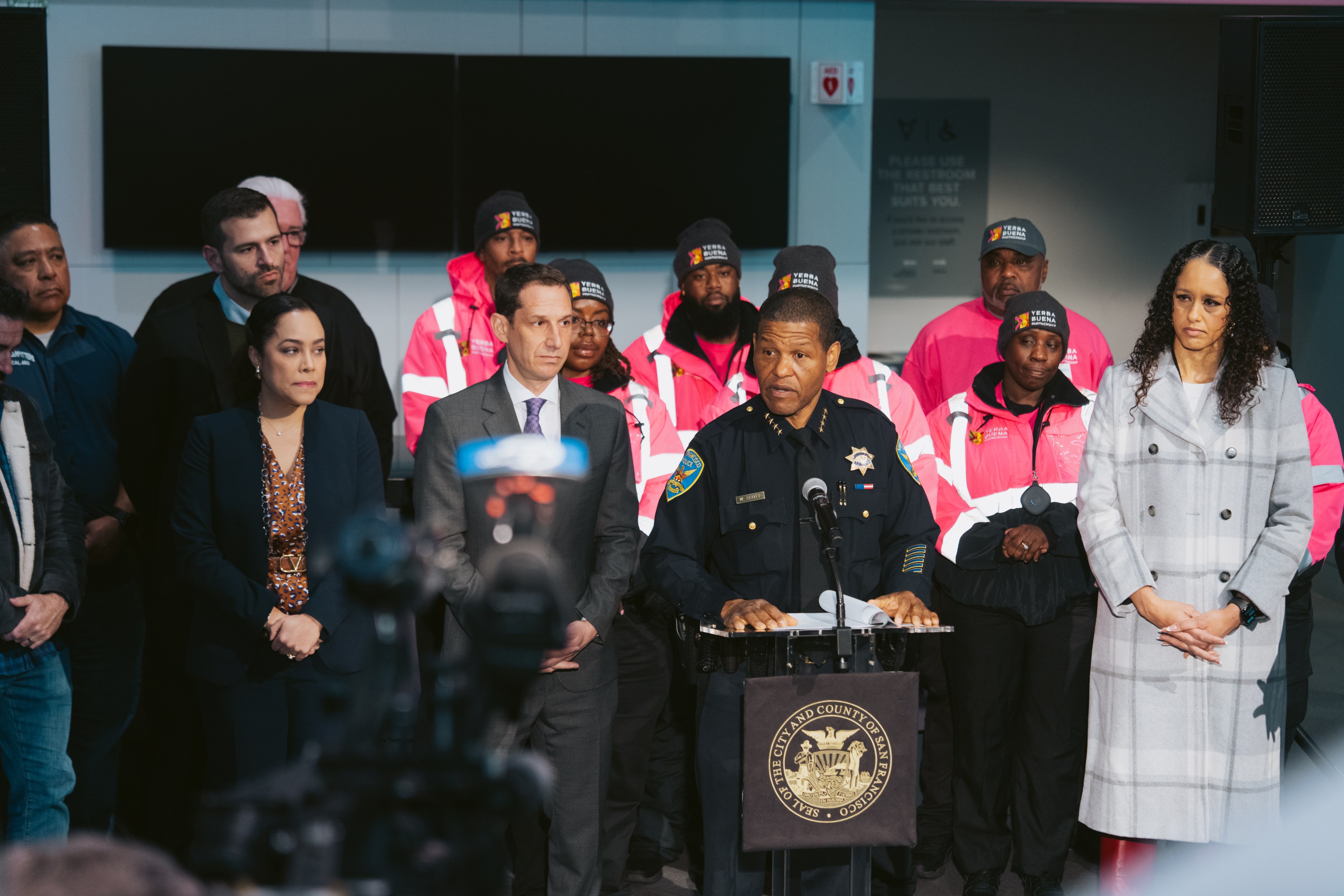 A police officer speaks at a podium with the City and County of San Francisco seal. Several people in suits and pink uniforms stand behind him, facing the audience.