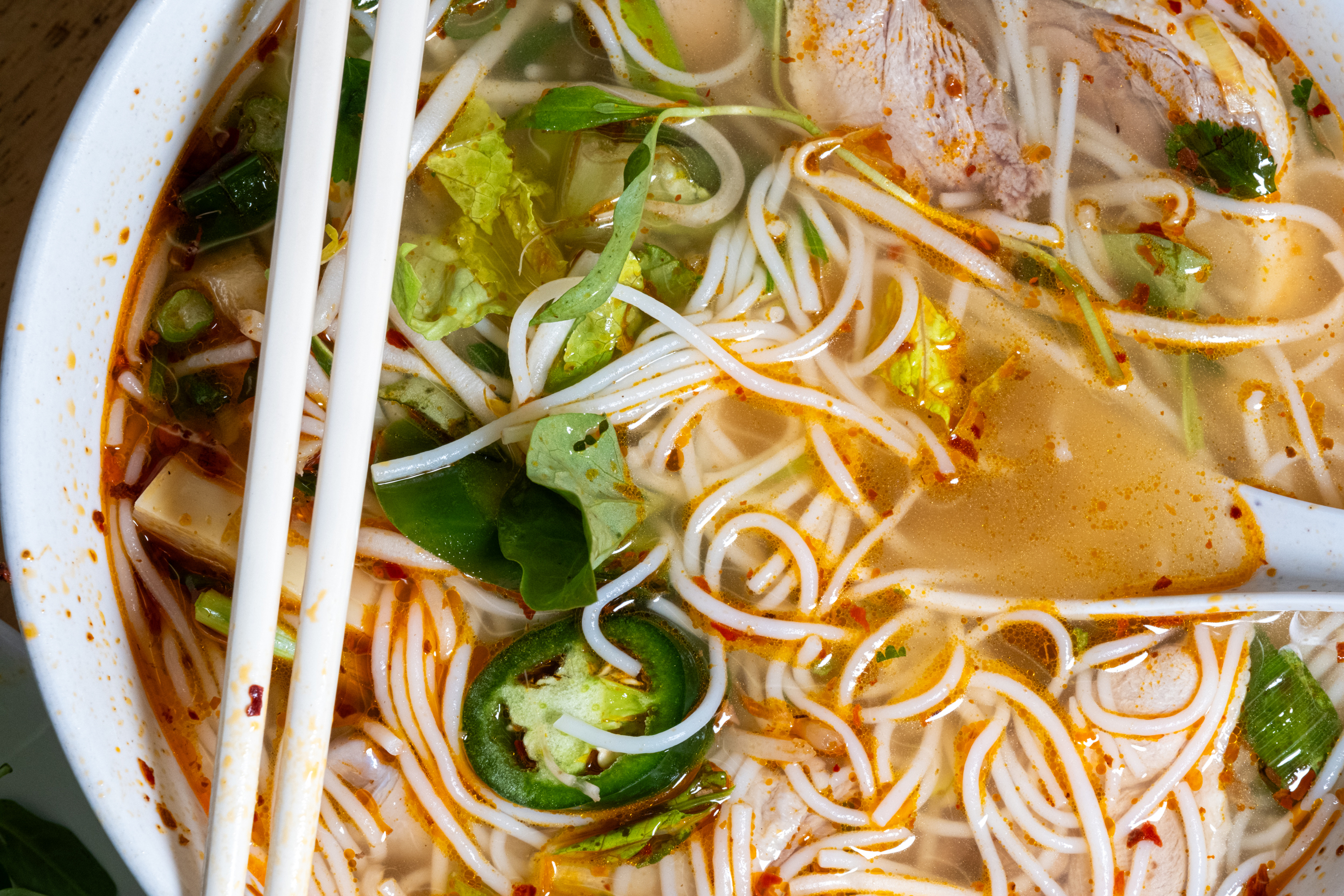 A bowl of noodle soup with rice noodles, sliced jalapeños, greens, and chunks of meat, accompanied by chopsticks and a spoon in a flavorful broth.