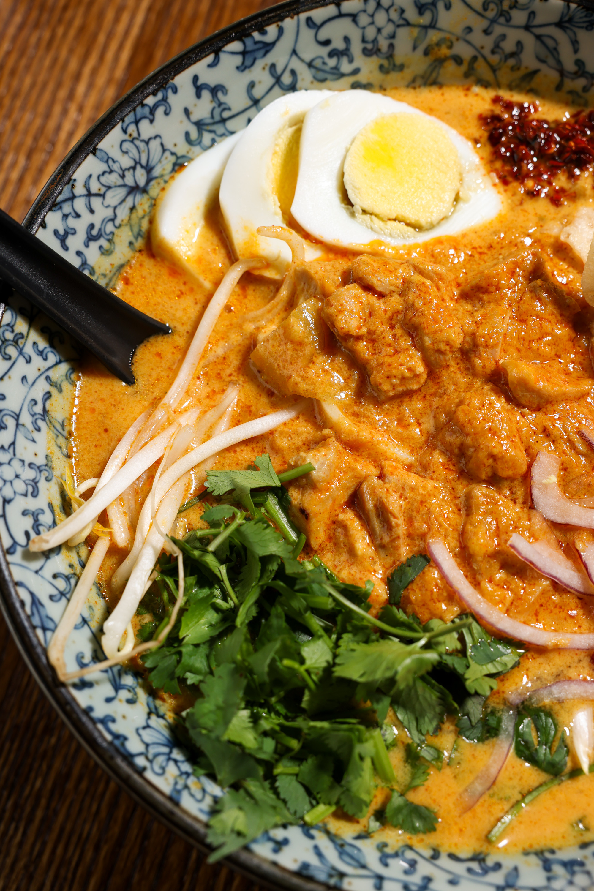 A bowl of spicy curry soup with chunks of meat, bean sprouts, boiled egg slices, fresh cilantro, and onions, served in a decorative blue-patterned bowl.