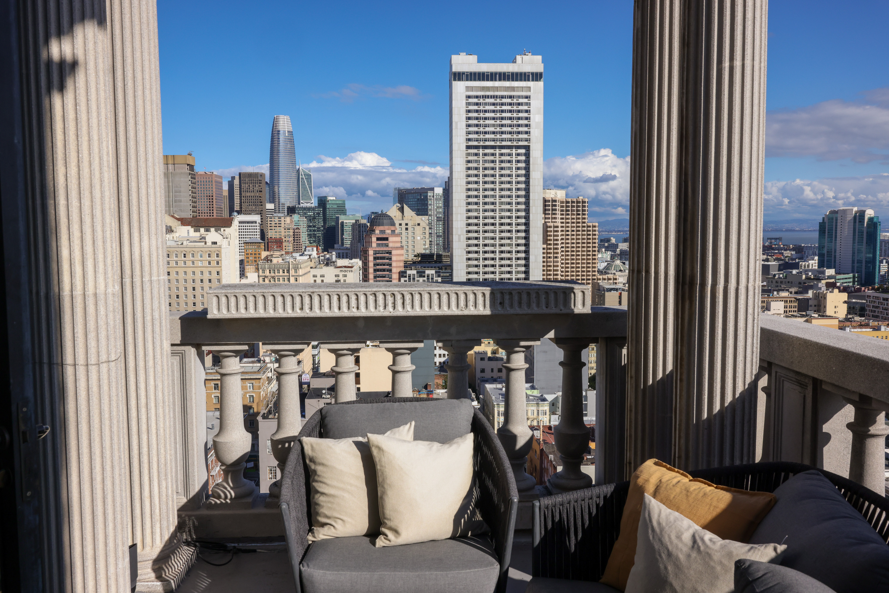 A rooftop terrace with elegant pillars offers a view of a city skyline featuring modern skyscrapers. Comfortable seating with cushions enhances the cozy setting.