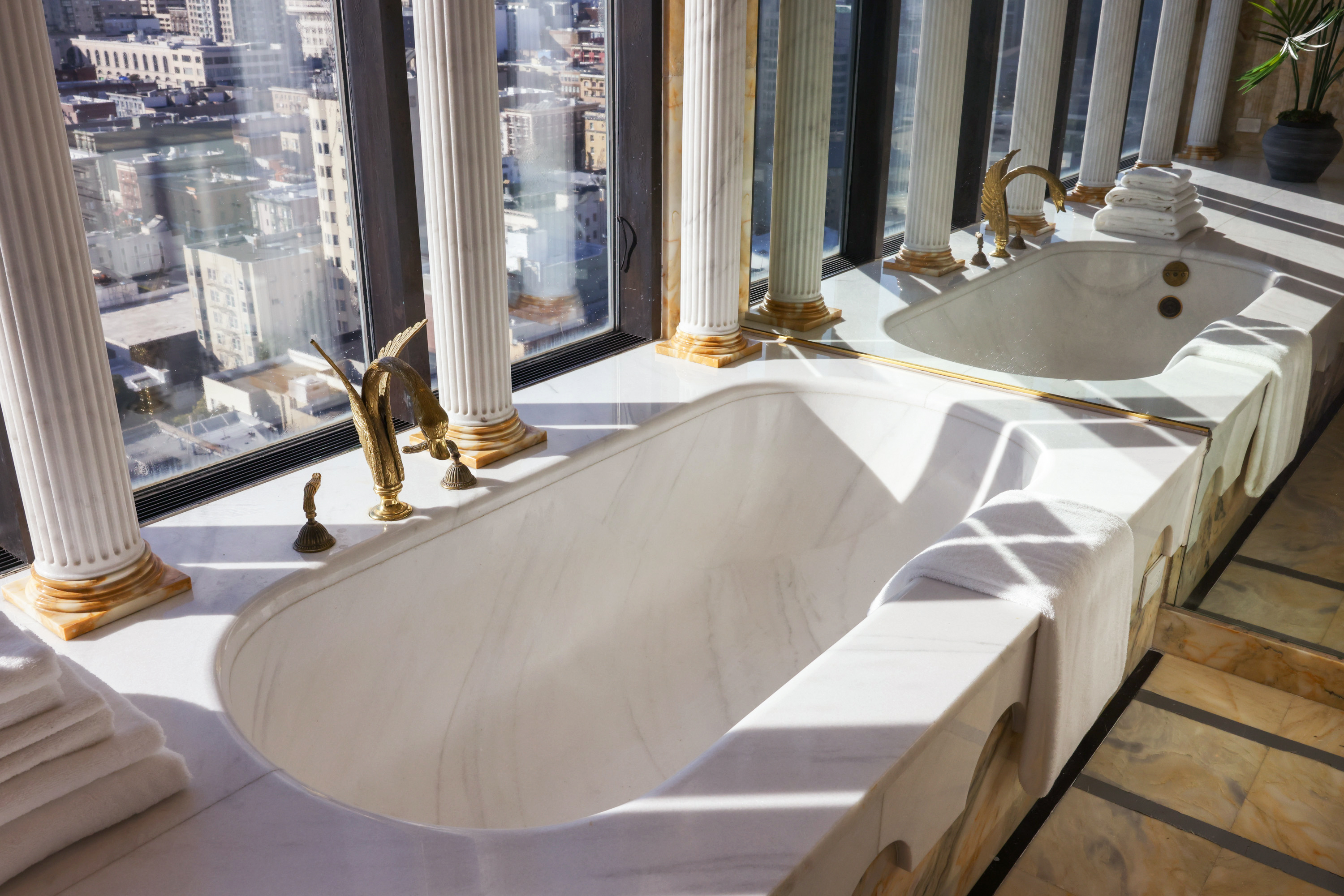 A luxurious marble bathtub with elegant gold fixtures is set beneath tall windows offering a city view. Tall white columns and neatly folded towels complete the scene.