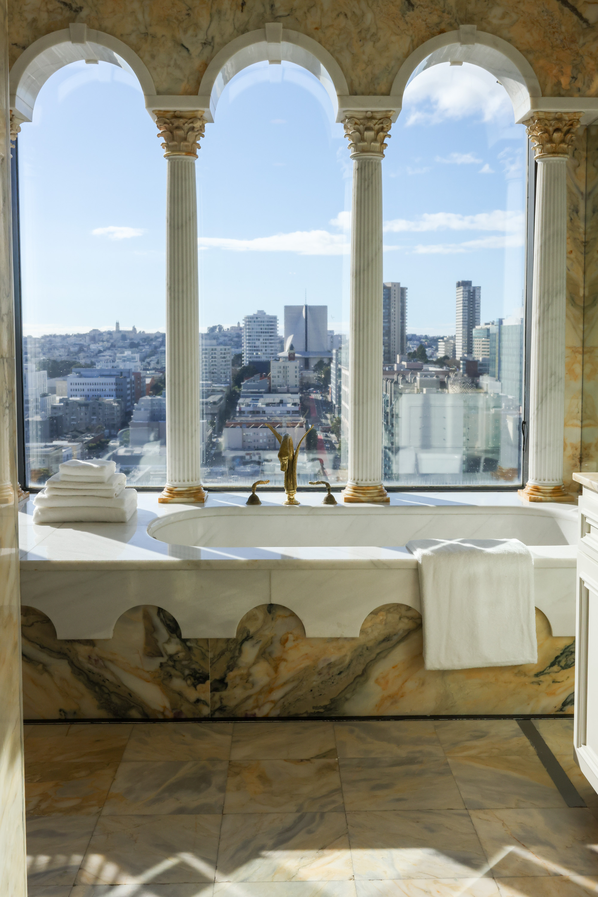 A luxurious marble bathroom with a bathtub overlooks a cityscape through large arched windows. Towels are neatly stacked nearby.