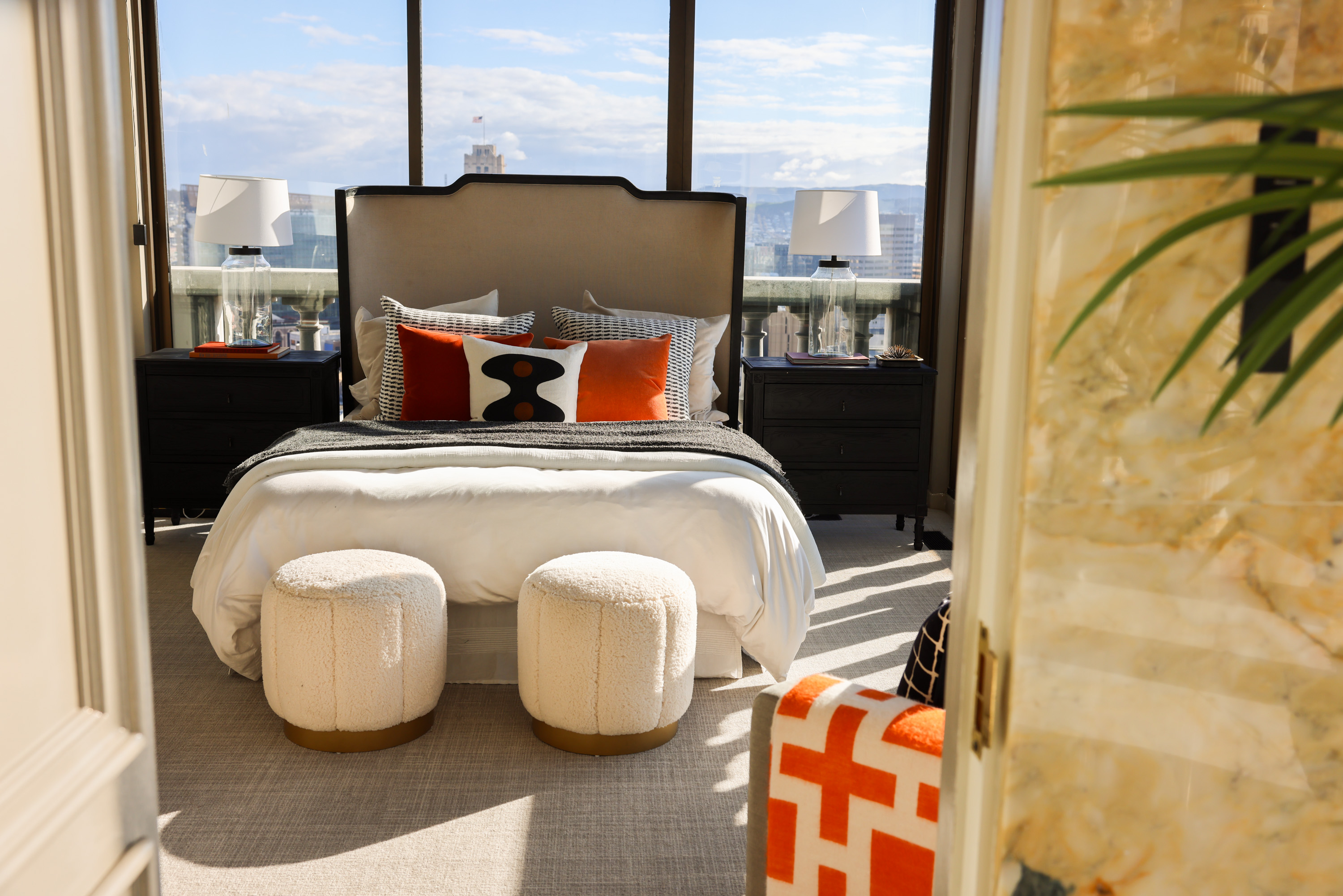 A cozy bedroom features a plush bed with decorative pillows, two round ottomans at the foot, nightstands with lamps, and a city view through large windows.