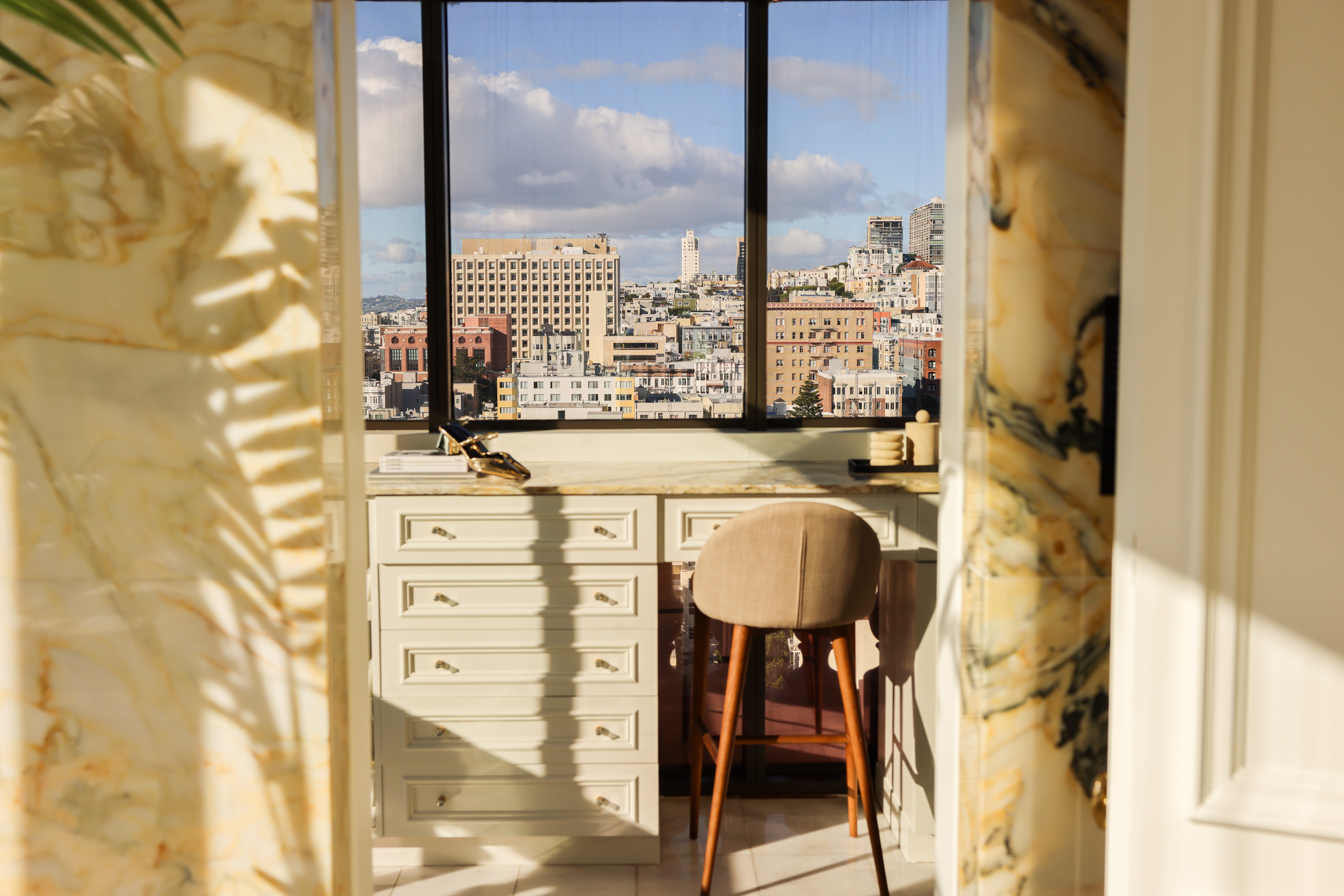 A sunlit room with a marble desk and a beige chair overlooks a cityscape through a large window, framed by marble walls.