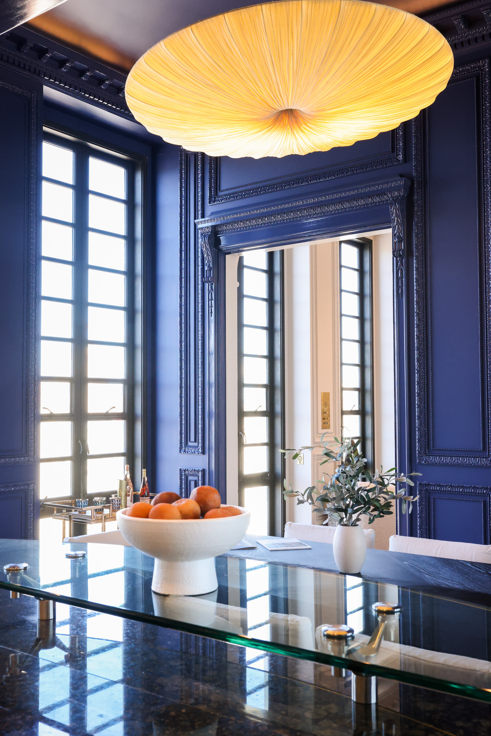 A modern room with dark blue walls features a large round ceiling light, a glass table, a white bowl of oranges, a small plant, and tall windows.