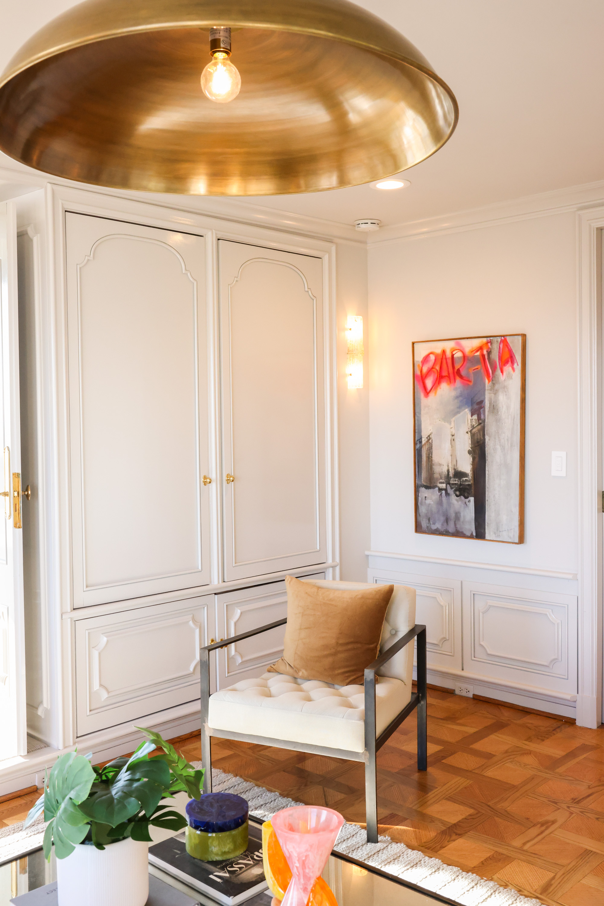 A cozy room features a cream armchair with a brown cushion, a large brass pendant light, abstract art with &quot;BARTA&quot; text, and a table with plants and decor.