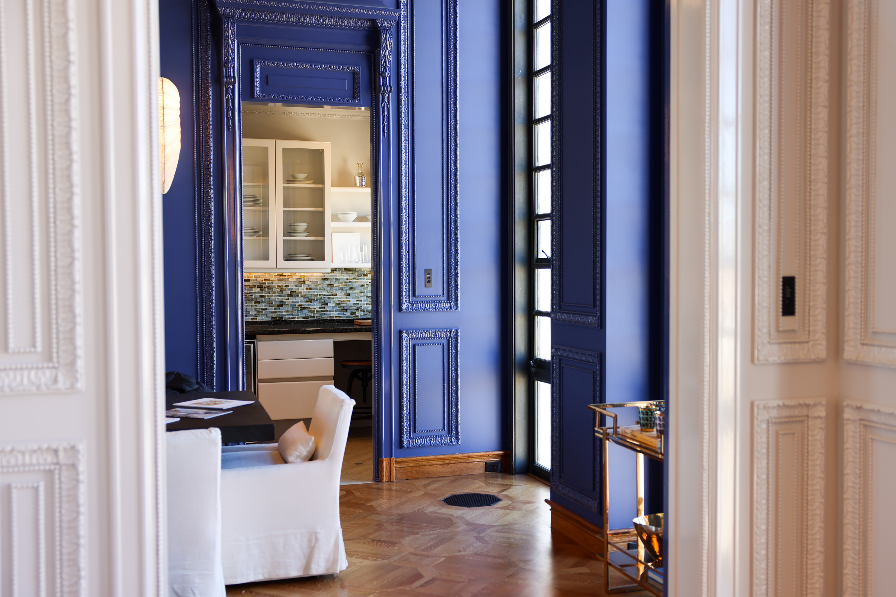 The image shows a lavish room with rich blue-paneled walls, a view into a kitchen with glass cabinets, and a dining area with white chairs and a wooden floor.