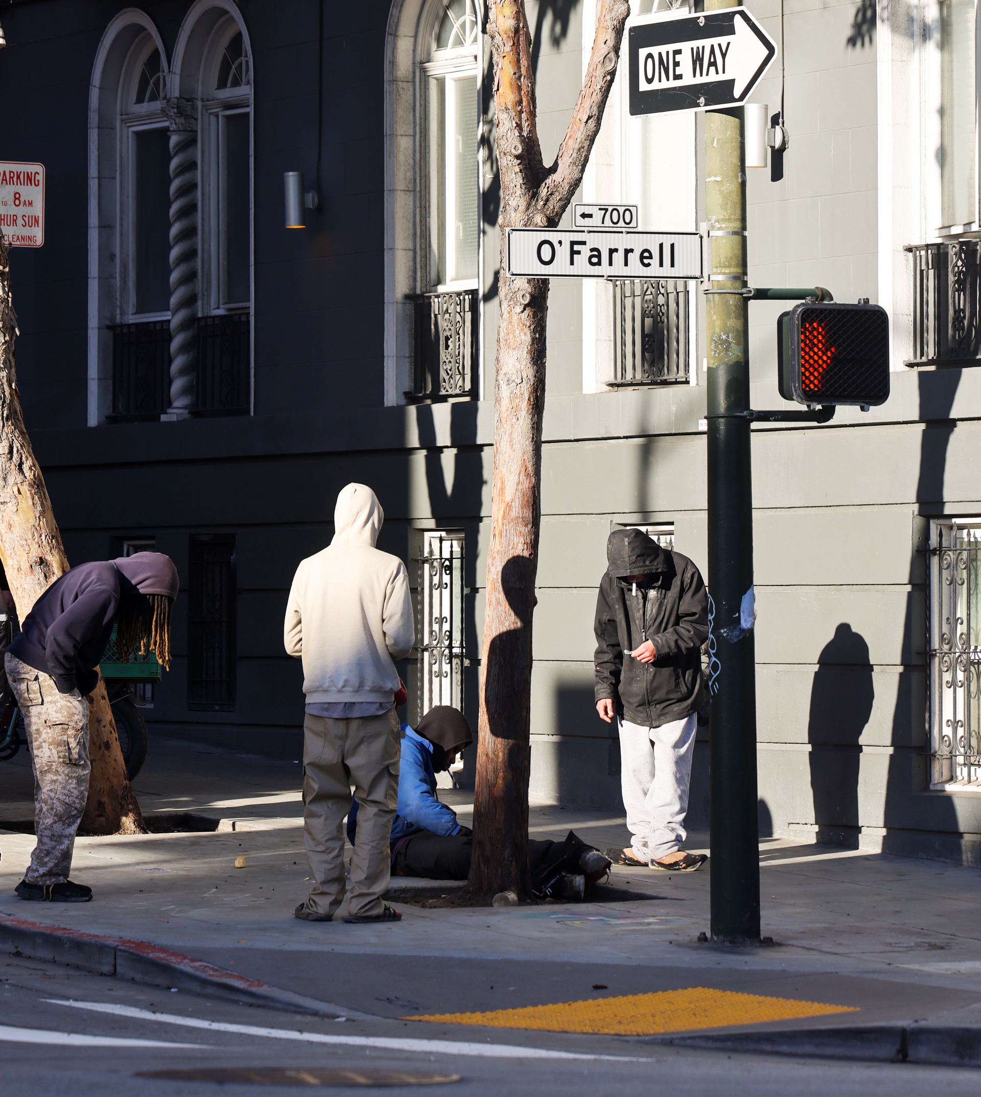 Four people are gathered on a city sidewalk near a street sign for O'Farrell. Two stand, one crouches, and one sits. A building and tree are in the background.