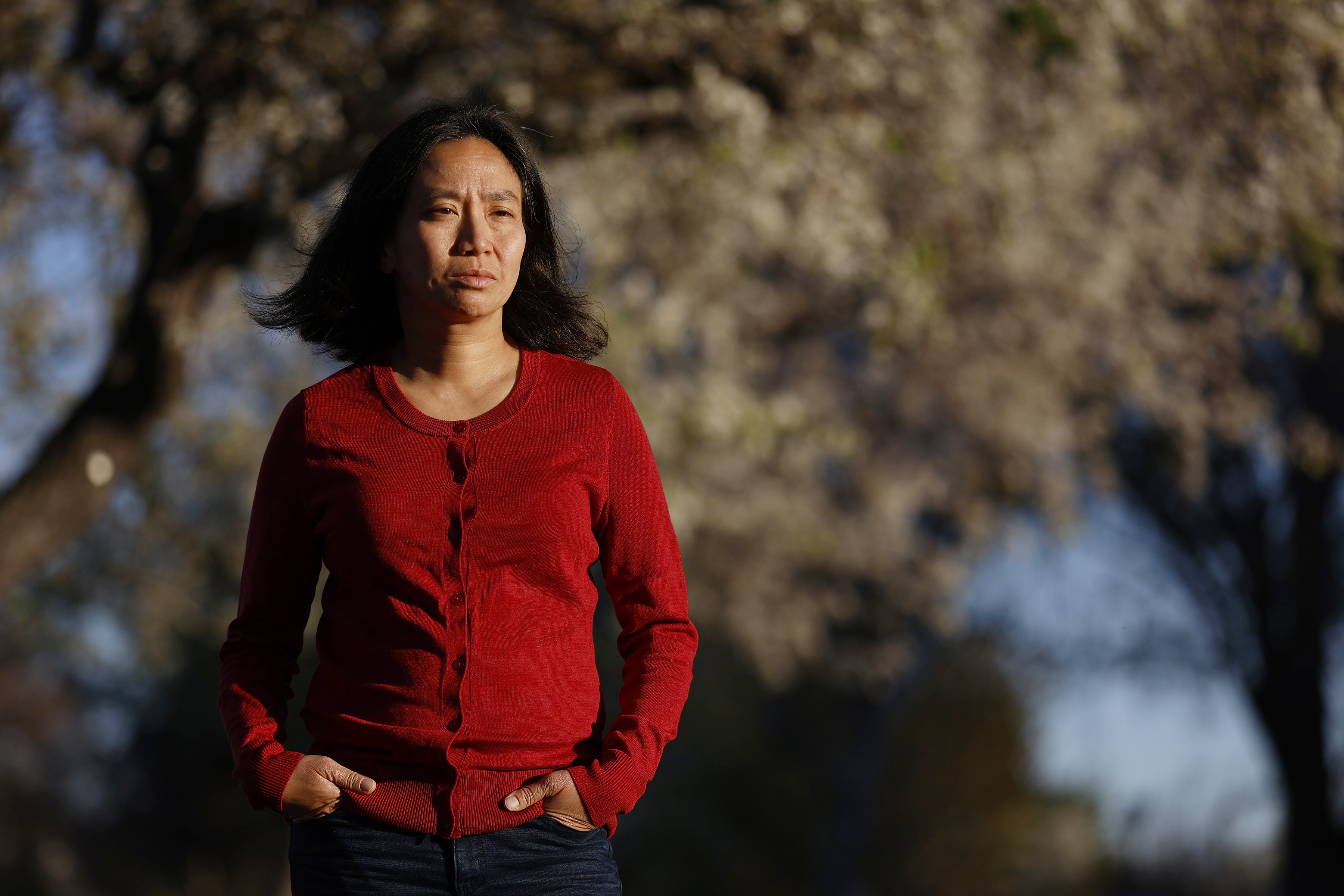 A person stands outdoors, wearing a red sweater and jeans, with hands in pockets. The background features blurred trees, suggesting a park setting.