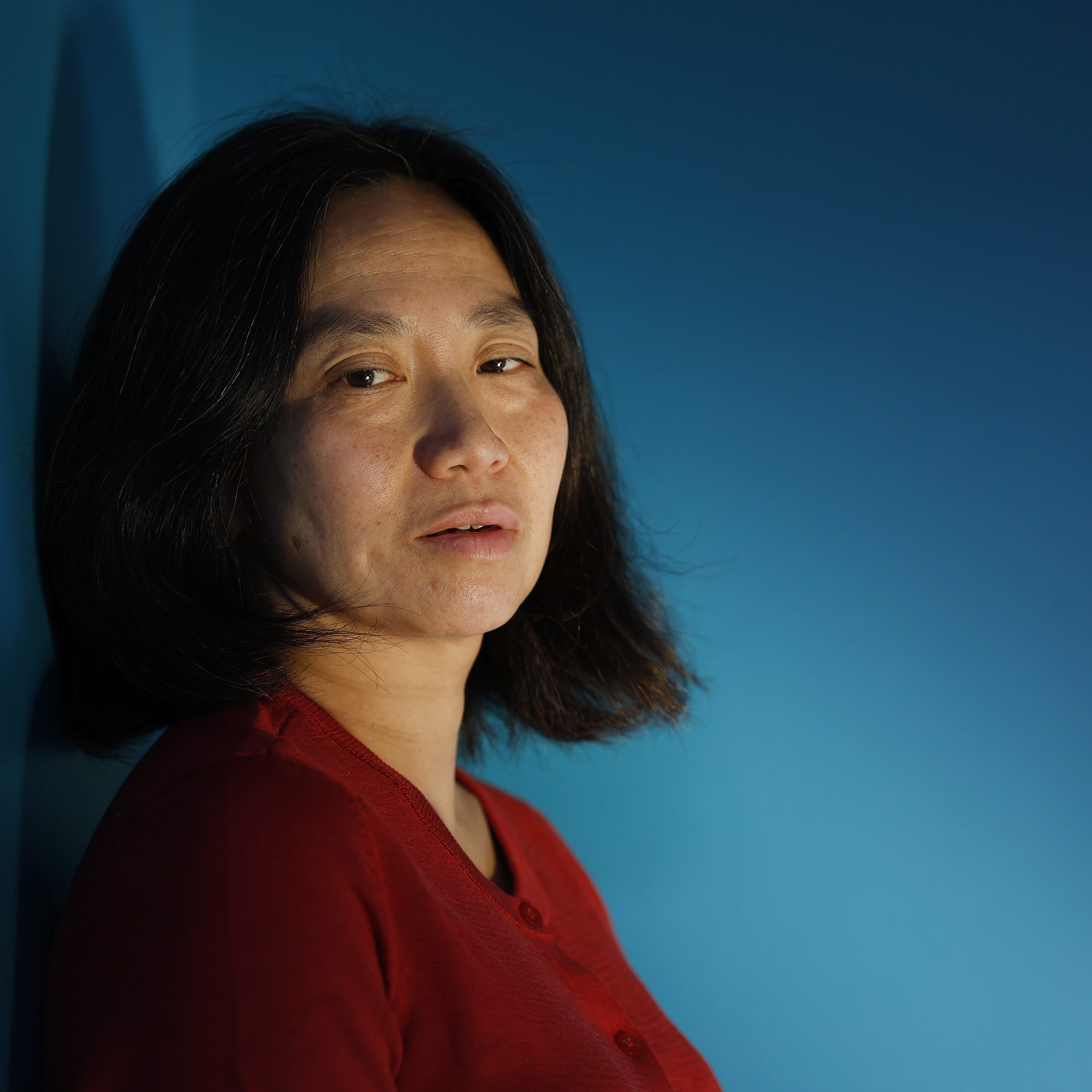 A person with shoulder-length dark hair, wearing a red top, stands against a blue background, looking directly at the camera with a neutral expression.