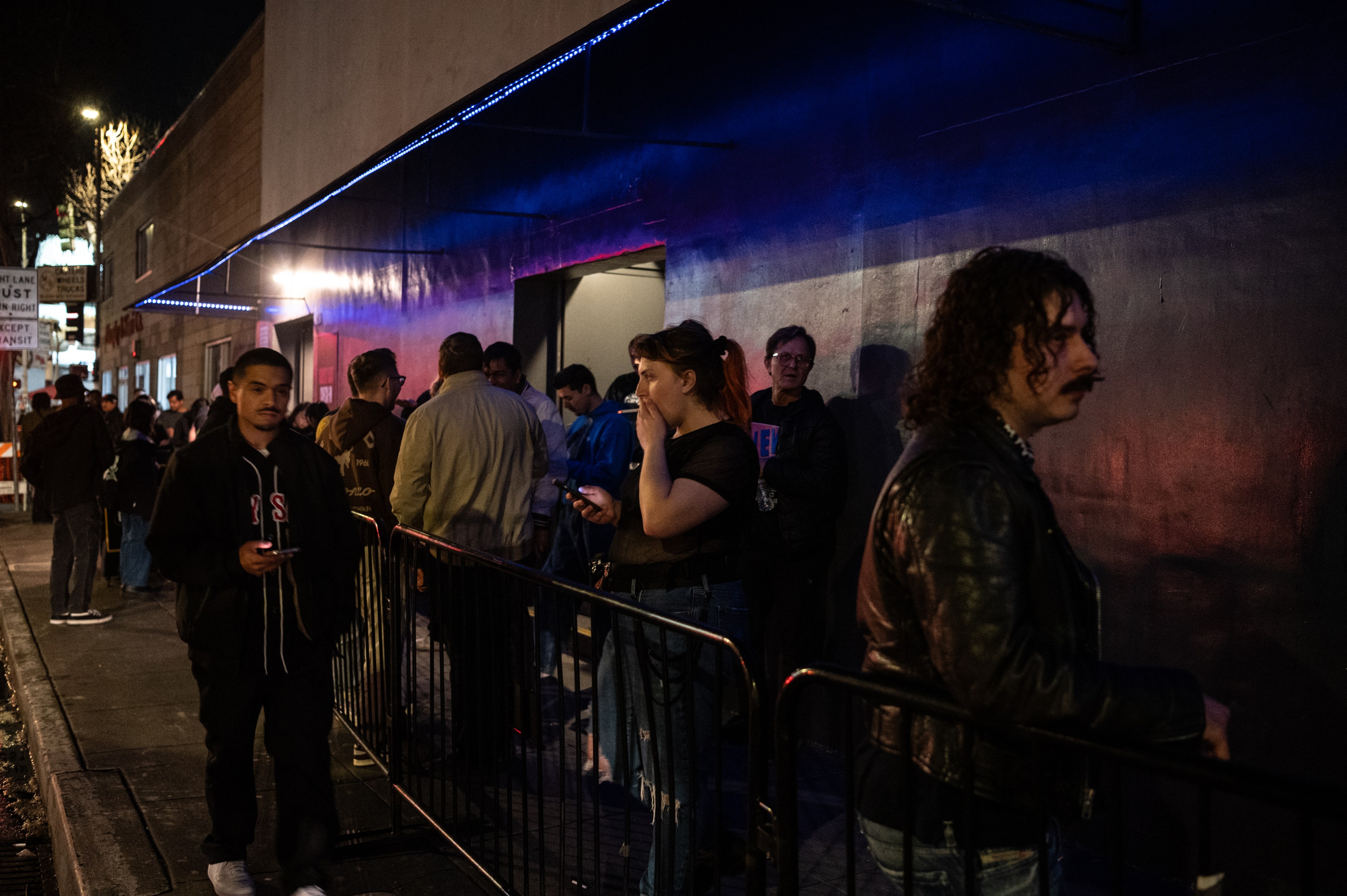 A group of people stand outside a building at night, illuminated by neon lights. Some are on their phones, and others are chatting, creating a lively atmosphere.