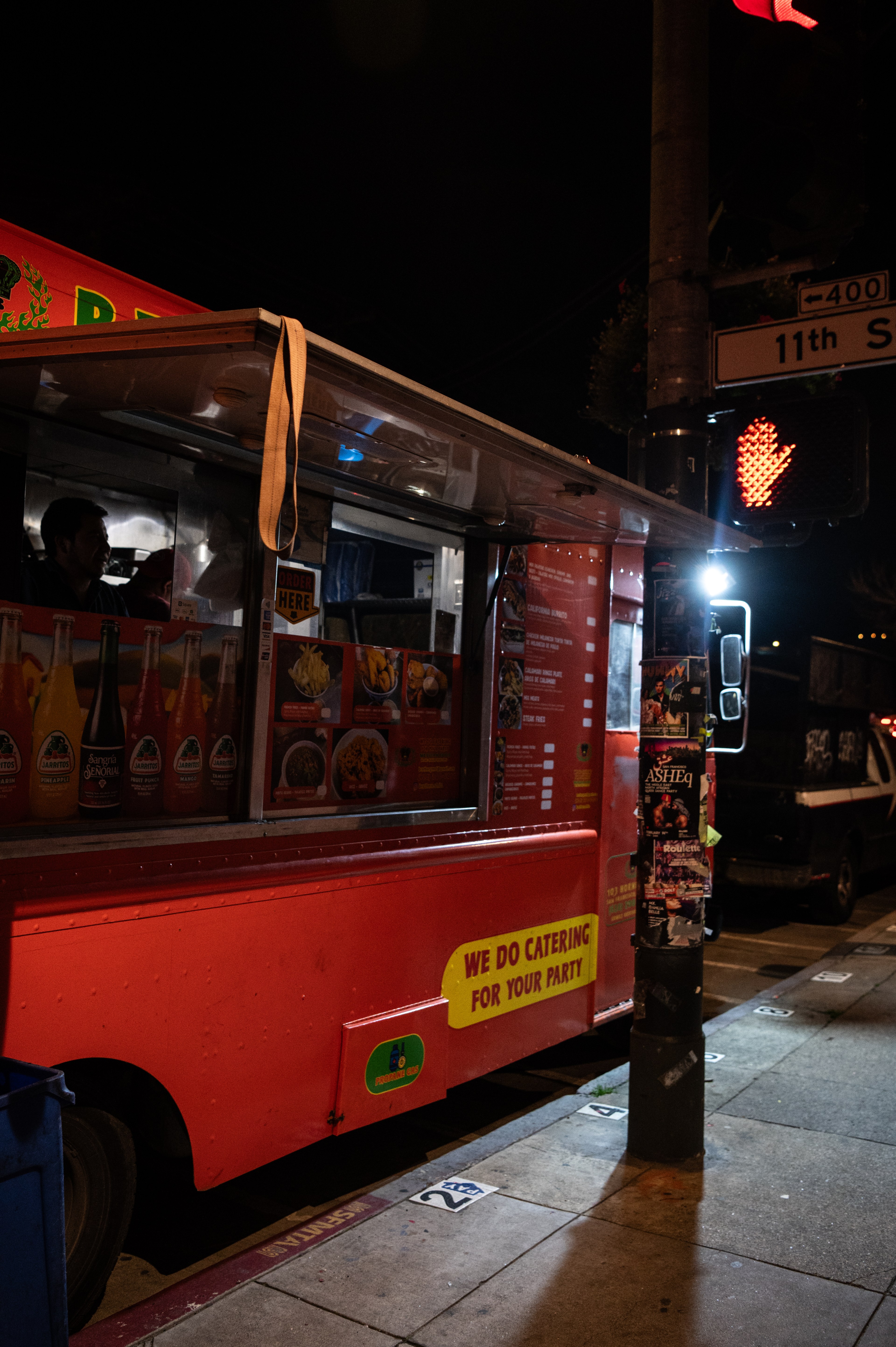 A brightly lit red food truck is parked at night, offering drinks and food. Menus and posters are visible, with a pedestrian &quot;don't walk&quot; signal illuminated nearby.