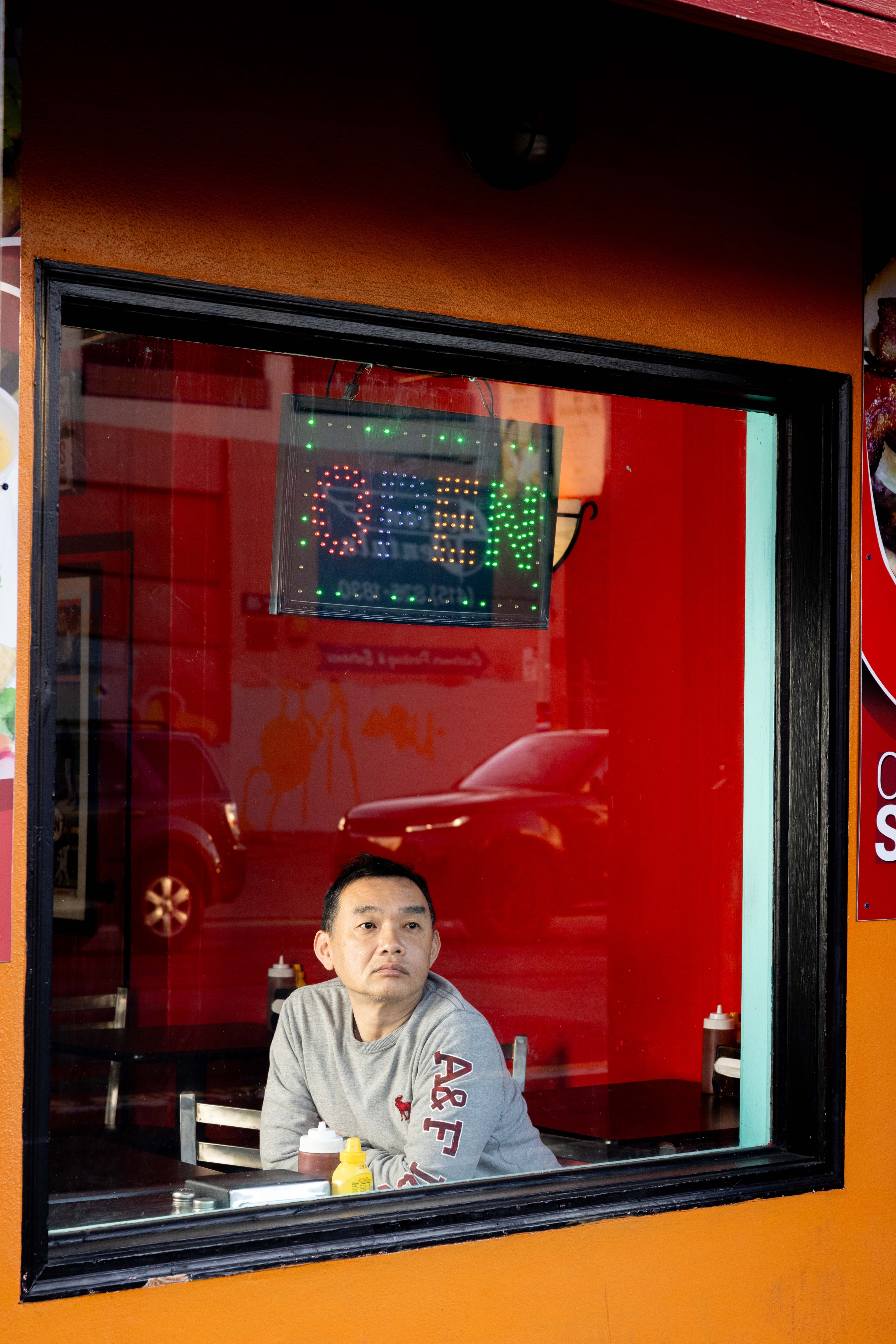 A person sits inside a café, gazing out the window. An &quot;OPEN&quot; sign glows above them. The interior is red with condiment bottles on the table.