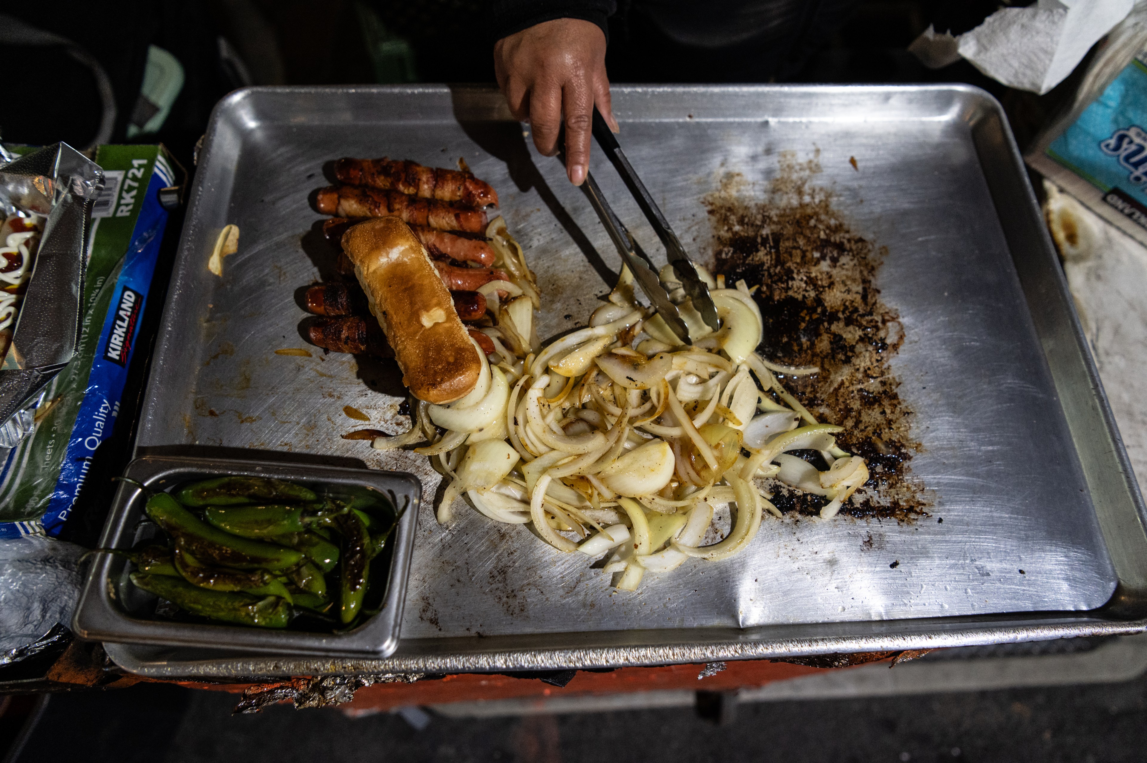 A hand uses tongs to cook onions on a metal griddle. Nearby, there are sausages wrapped in bacon and a toasted bun. A container of green peppers is also visible.