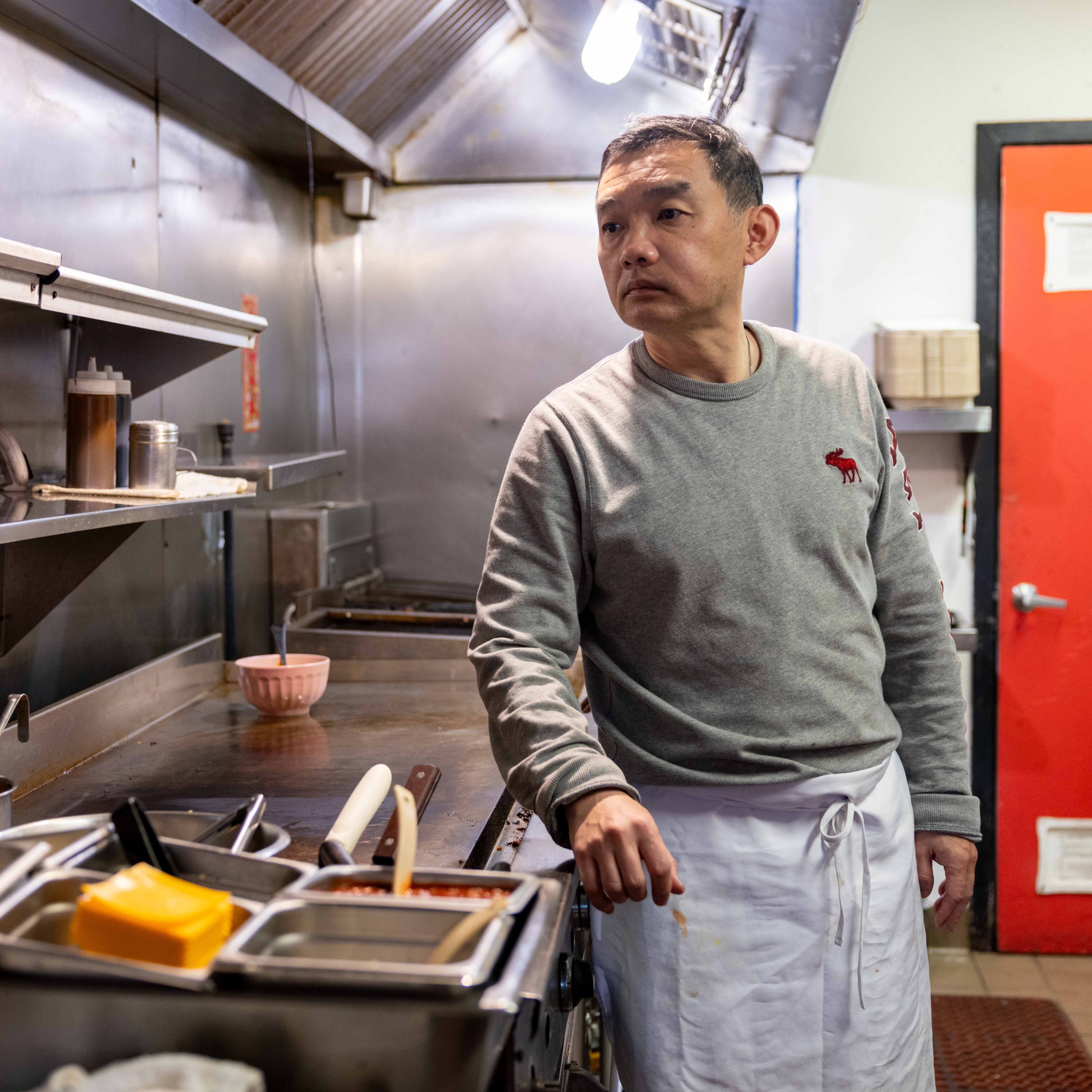 A man in a gray sweater and white apron stands in a commercial kitchen. He looks to the side, with utensils and kitchen equipment around him. A red door is visible.