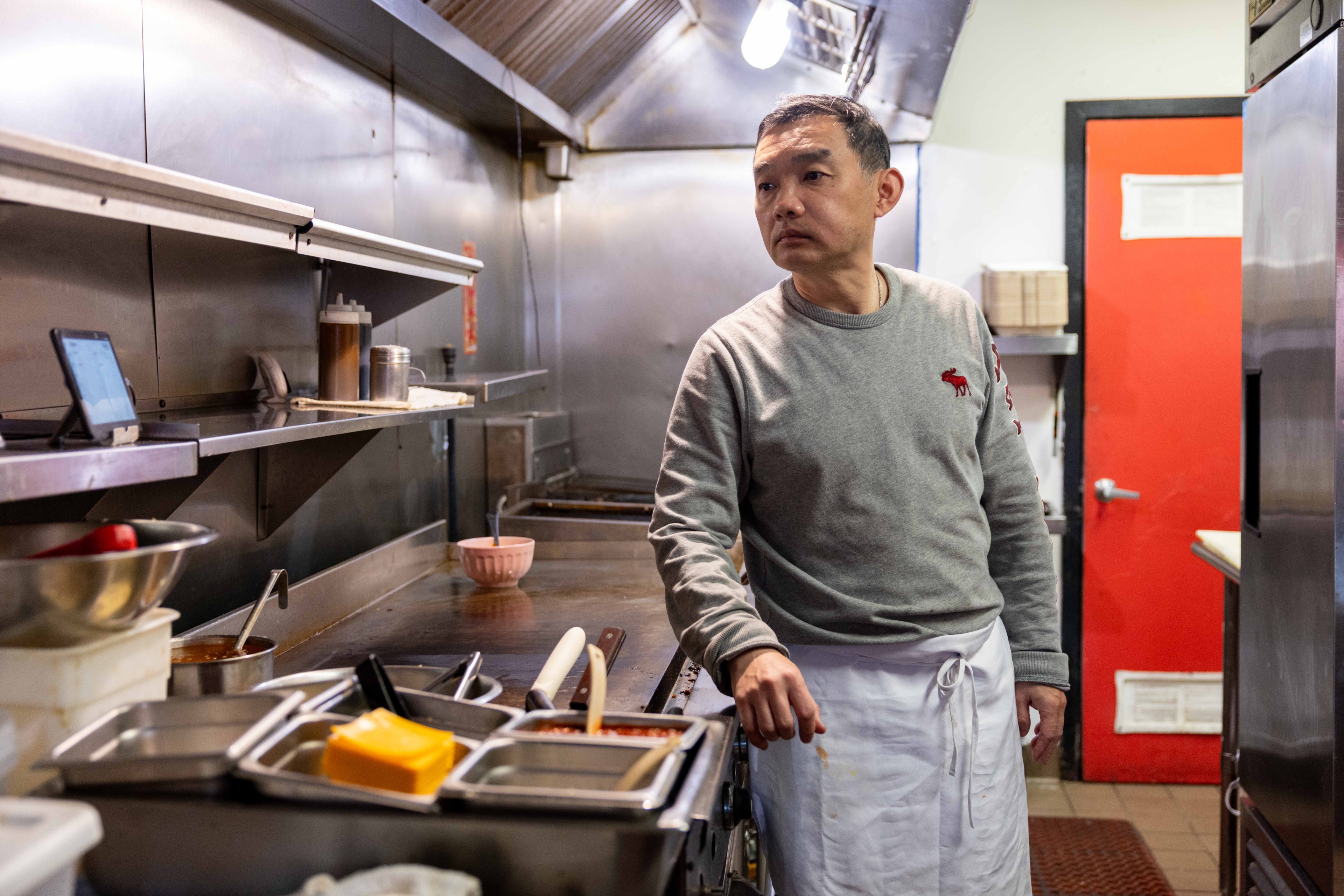 A man in a gray sweater and white apron stands in a commercial kitchen. He looks to the side, with utensils and kitchen equipment around him. A red door is visible.