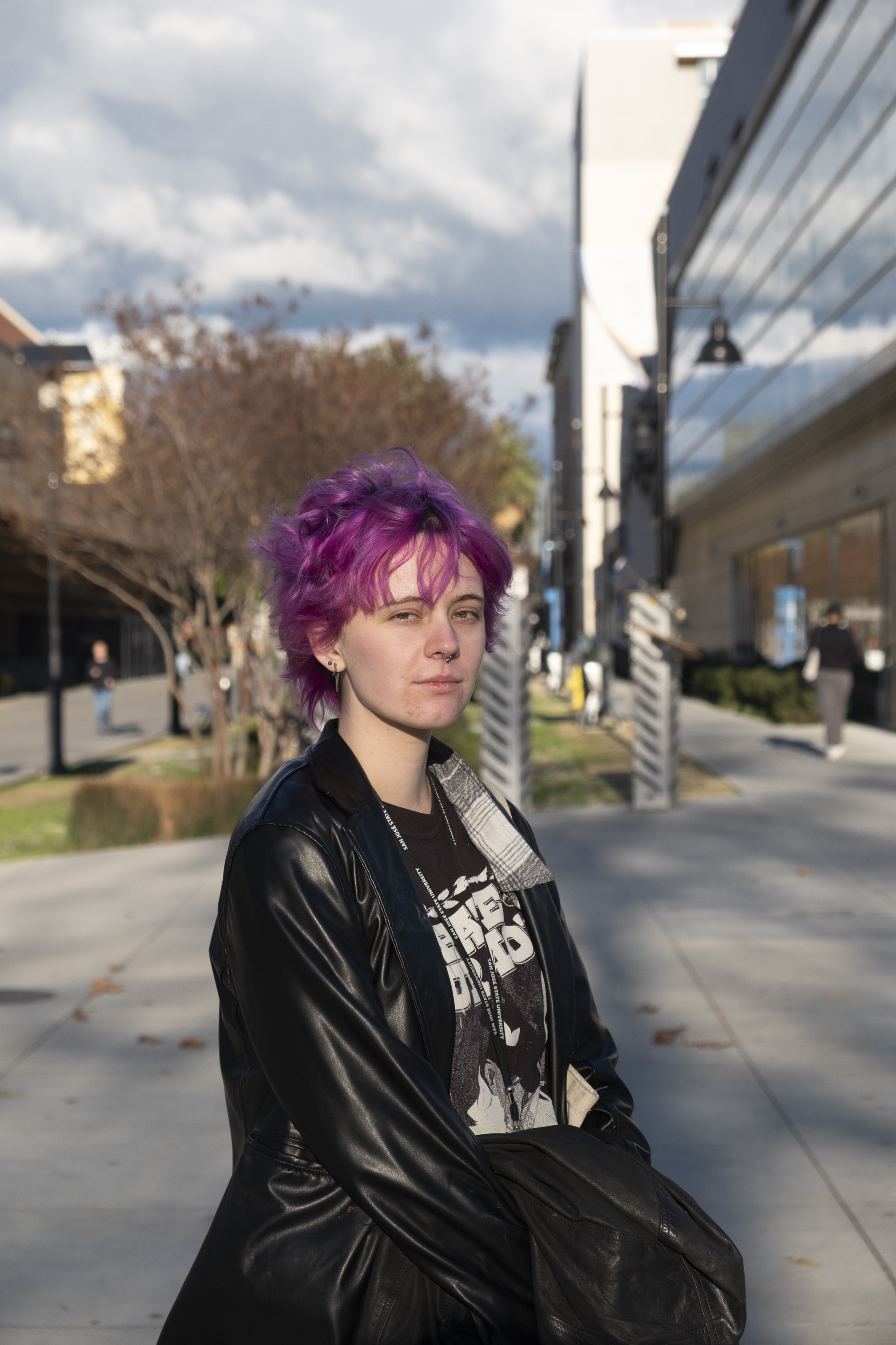 A person with bright pink hair stands on a sidewalk, wearing a black leather jacket and a graphic tee. The background includes buildings and trees under a cloudy sky.