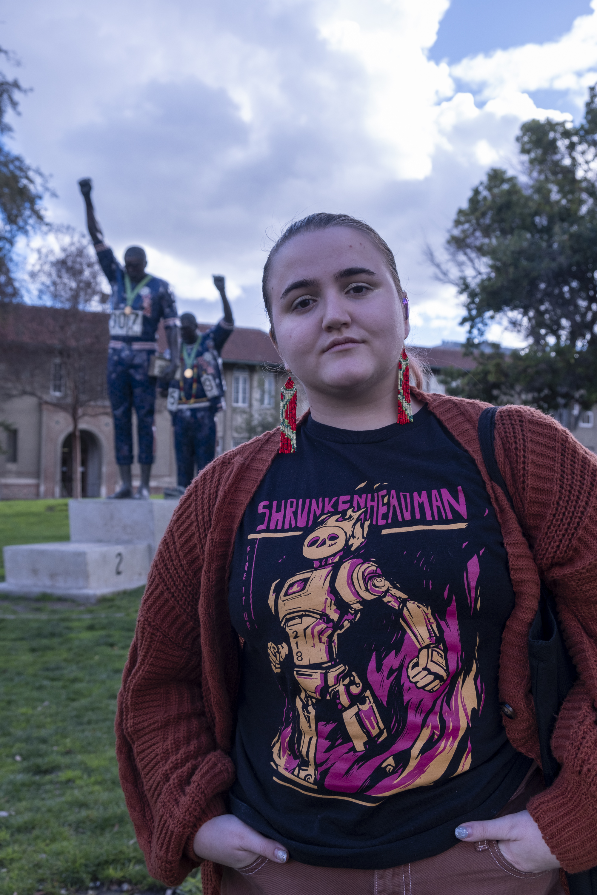 A person stands in the foreground wearing a graphic t-shirt and red cardigan. Behind them is a statue of two figures with raised fists, set against a cloudy sky.