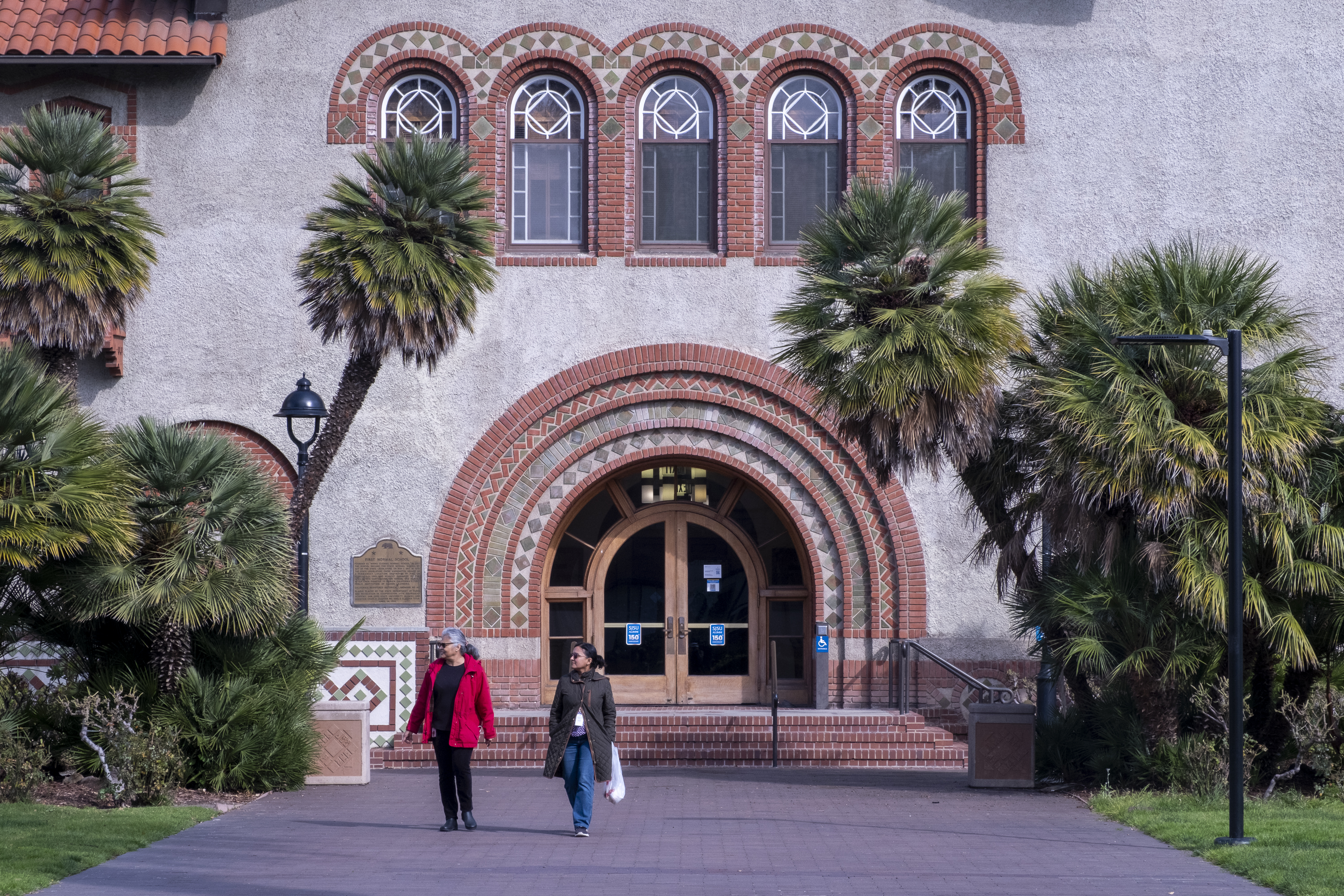 Two people walk on a pathway in front of a historic building with arched windows and ornate brickwork, surrounded by palm trees and a grassy area.