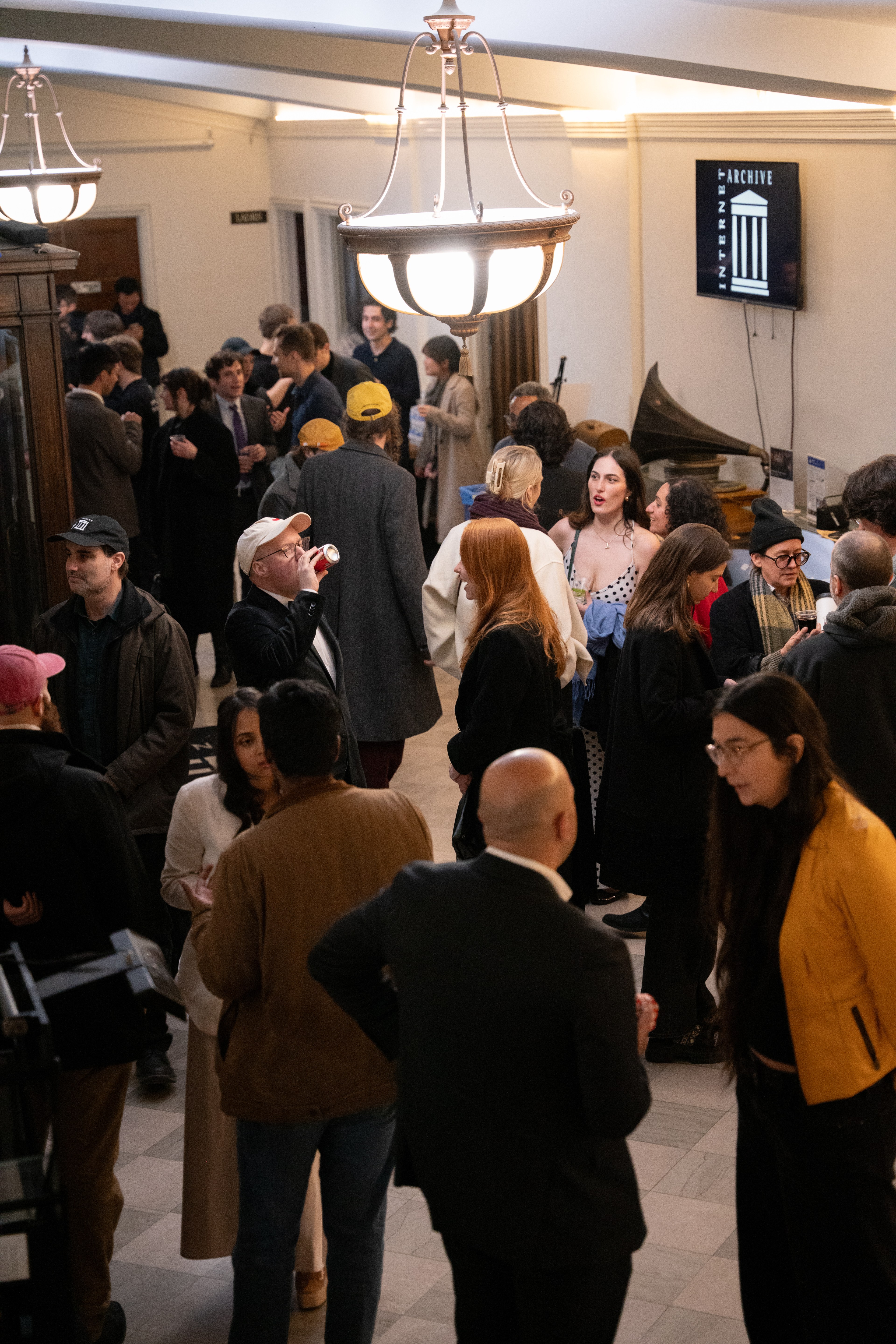 A lively indoor gathering with people socializing, some holding drinks. A large light fixture hangs from the ceiling, and a sign reads &quot;Internet Archive.&quot;