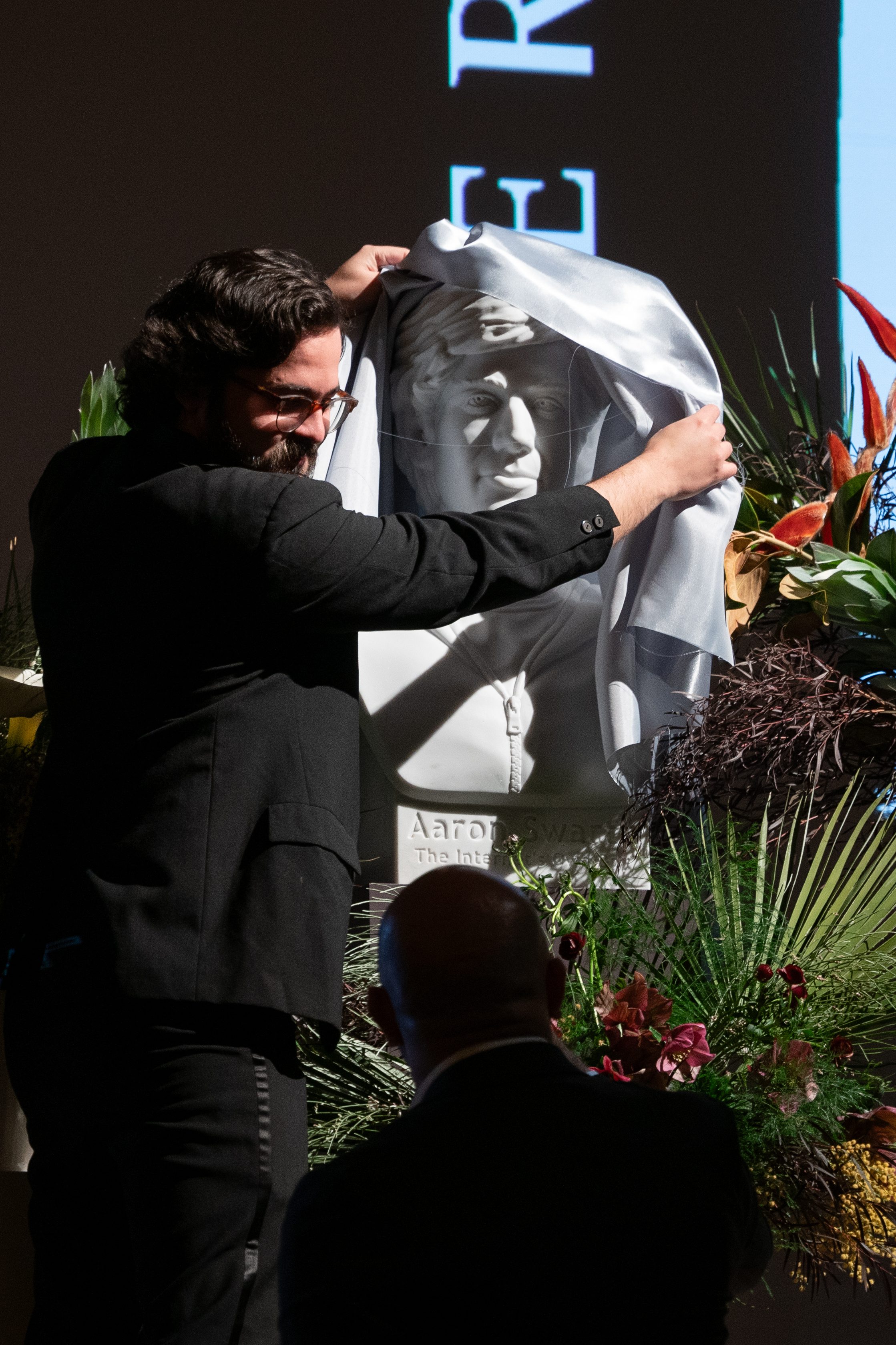 A man unveils a draped bust surrounded by lush greenery and flowers. The background features a minimalist building illustration.