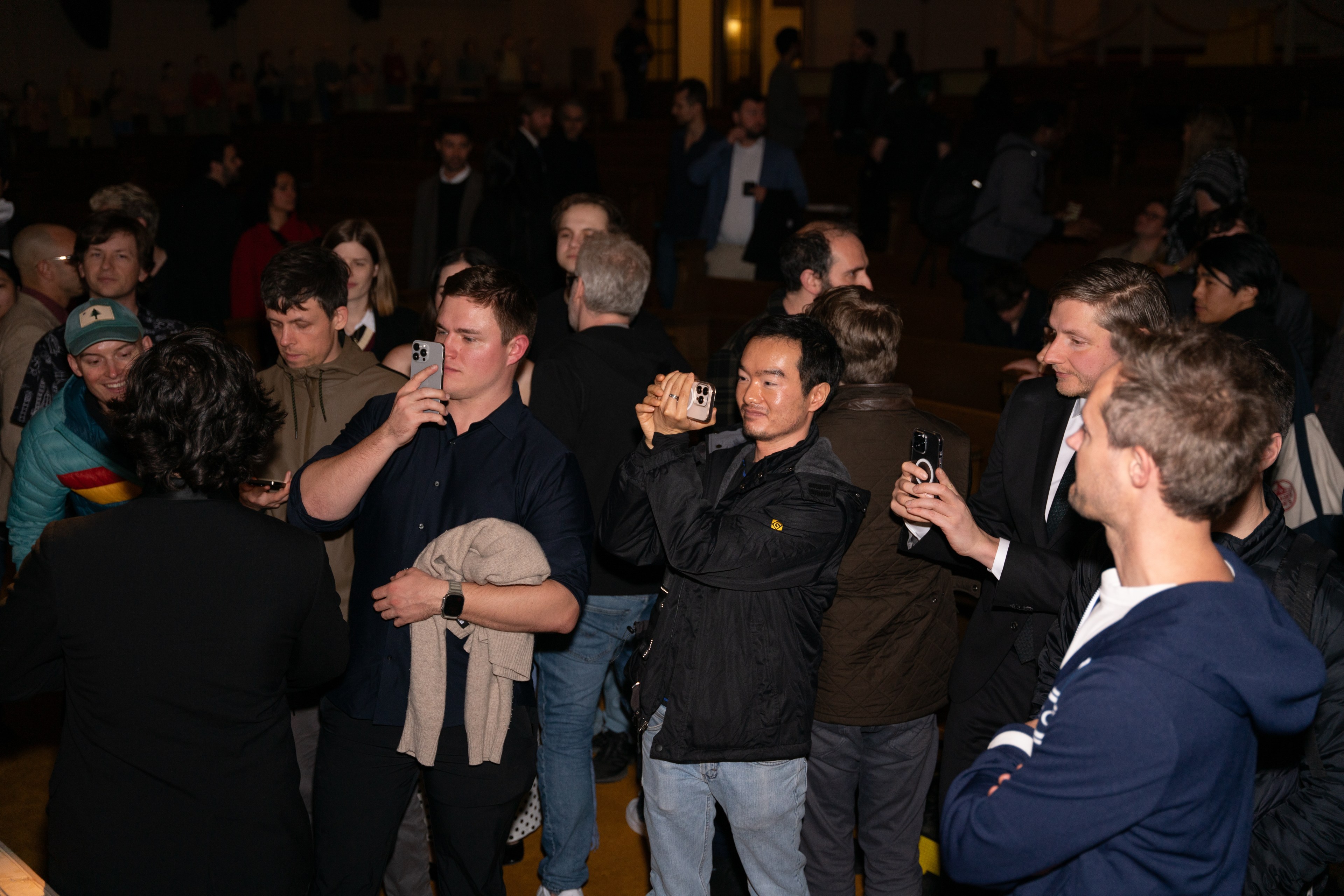 A group of people in a dimly lit room are holding phones, seemingly taking photos or videos. The atmosphere appears lively and focused.