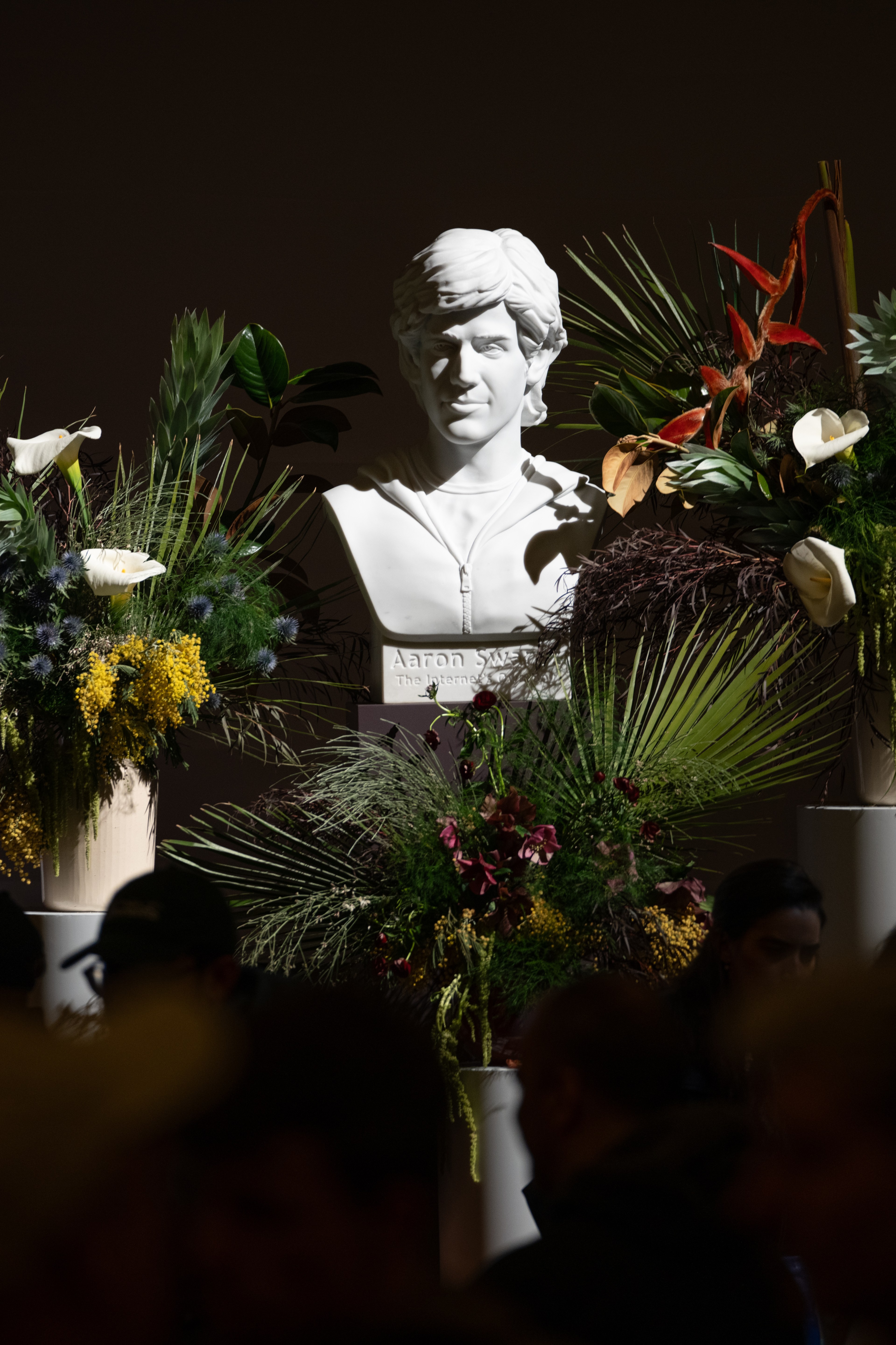 A white bust is surrounded by lush flower arrangements featuring palms, calla lilies, and orange accents. It's set against a dark background with silhouetted people in the foreground.