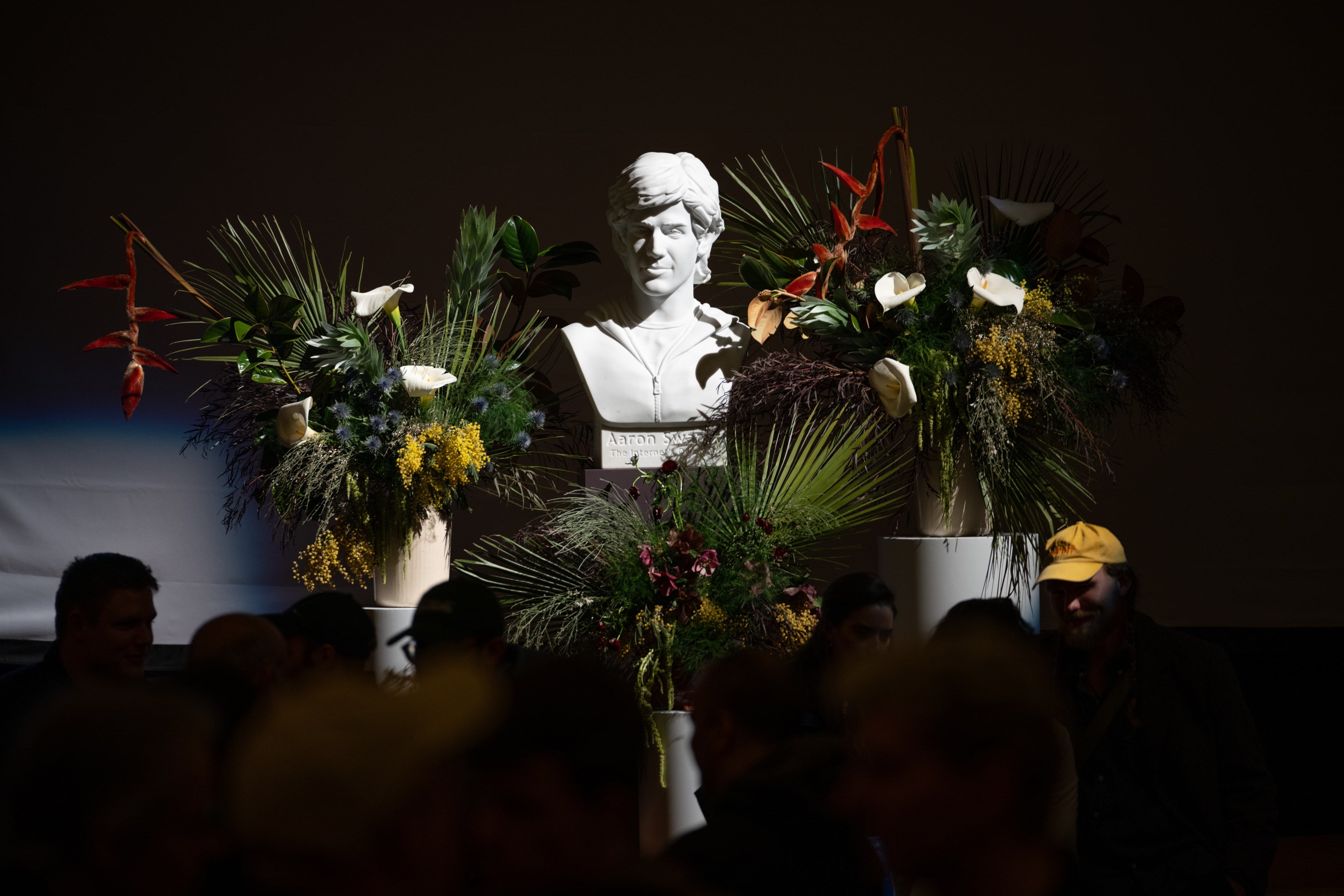 A white bust is surrounded by lush flower arrangements featuring palms, calla lilies, and orange accents. It's set against a dark background with silhouetted people in the foreground.