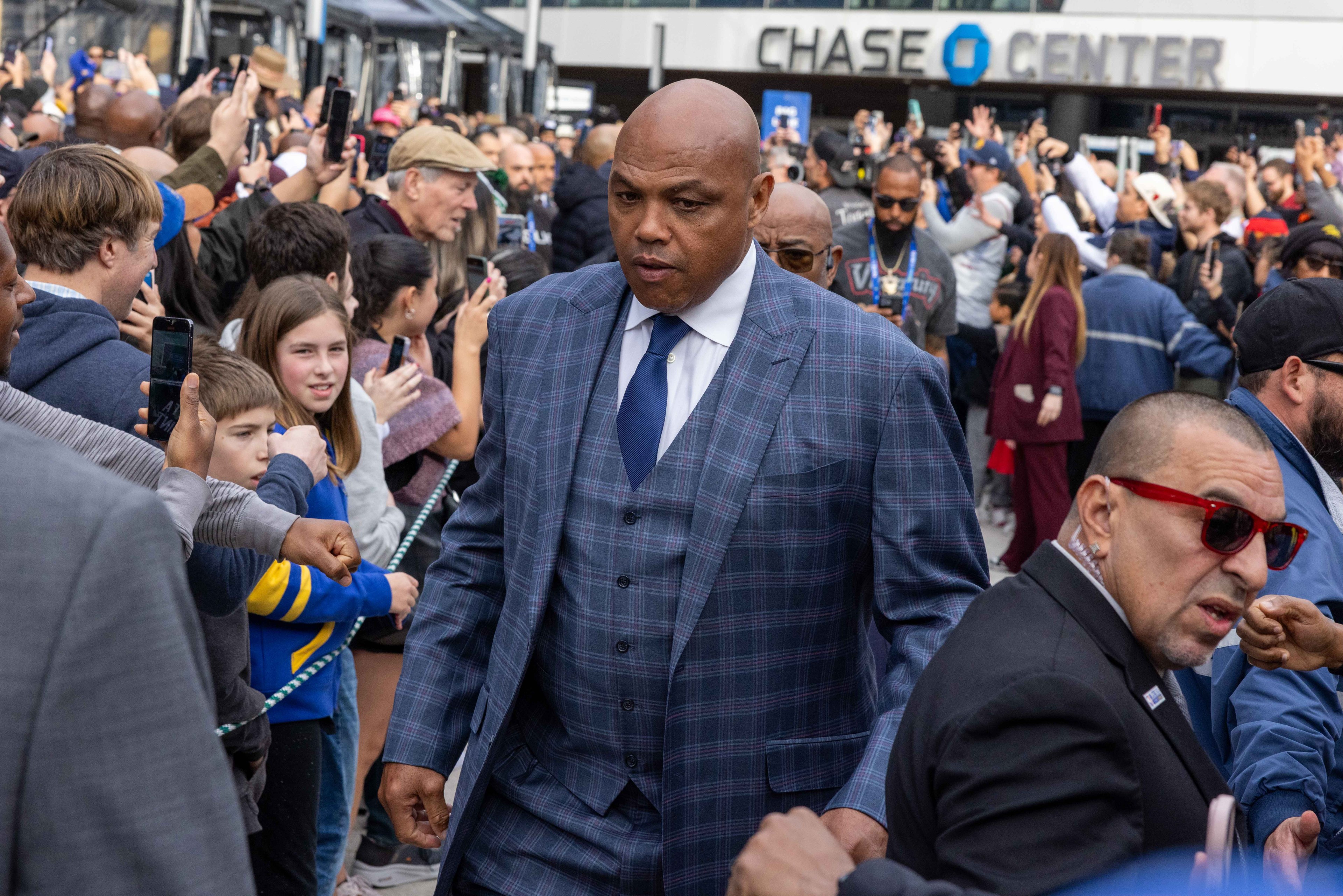 A man in a plaid suit, surrounded by a large crowd taking photos, walks outside a building named "Chase Center." People eagerly reach out around him.