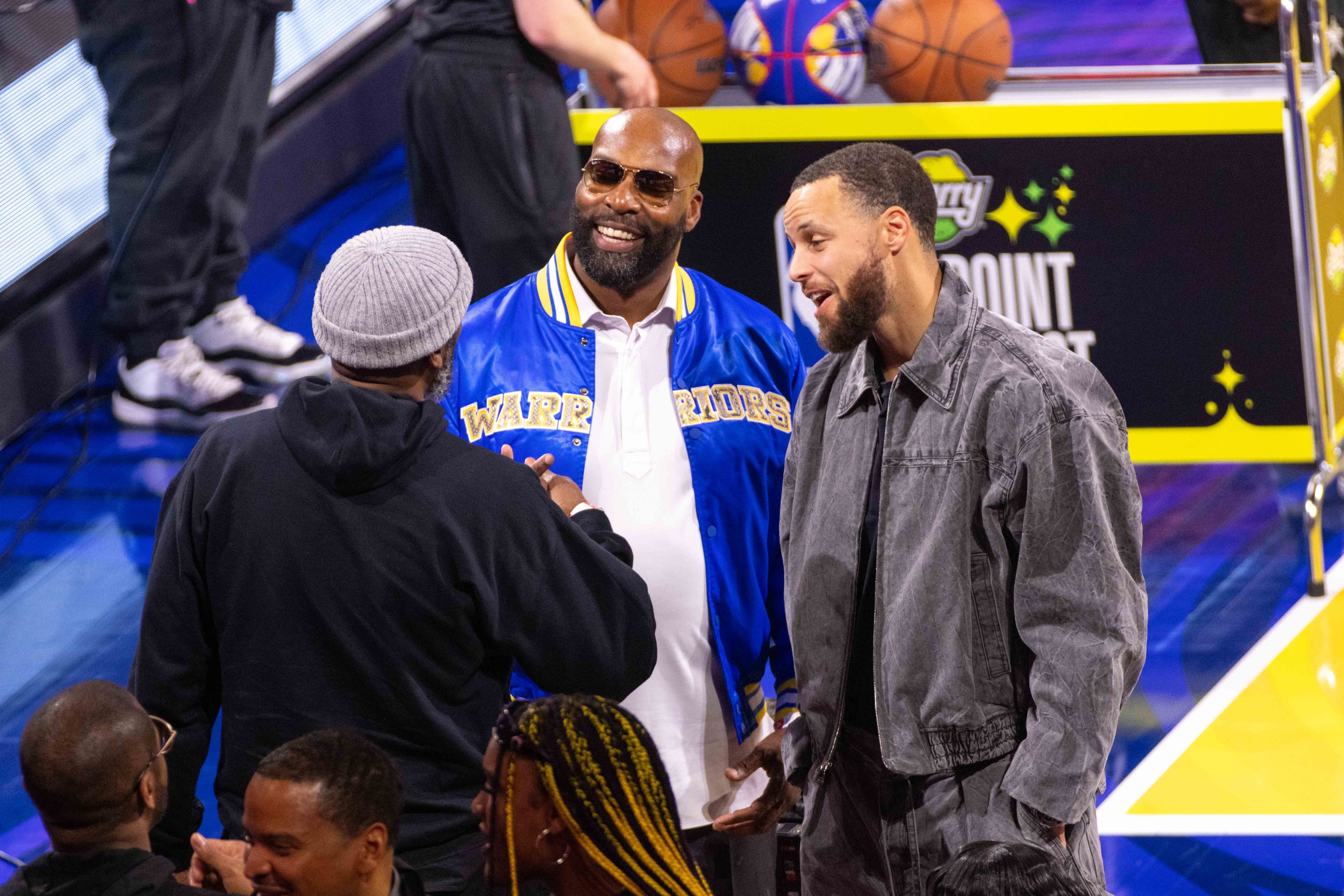 A group of men are talking in a sports arena setting. One man is wearing a blue &quot;Warriors&quot; jacket, while another is in a gray outfit. They appear to be having a friendly conversation.