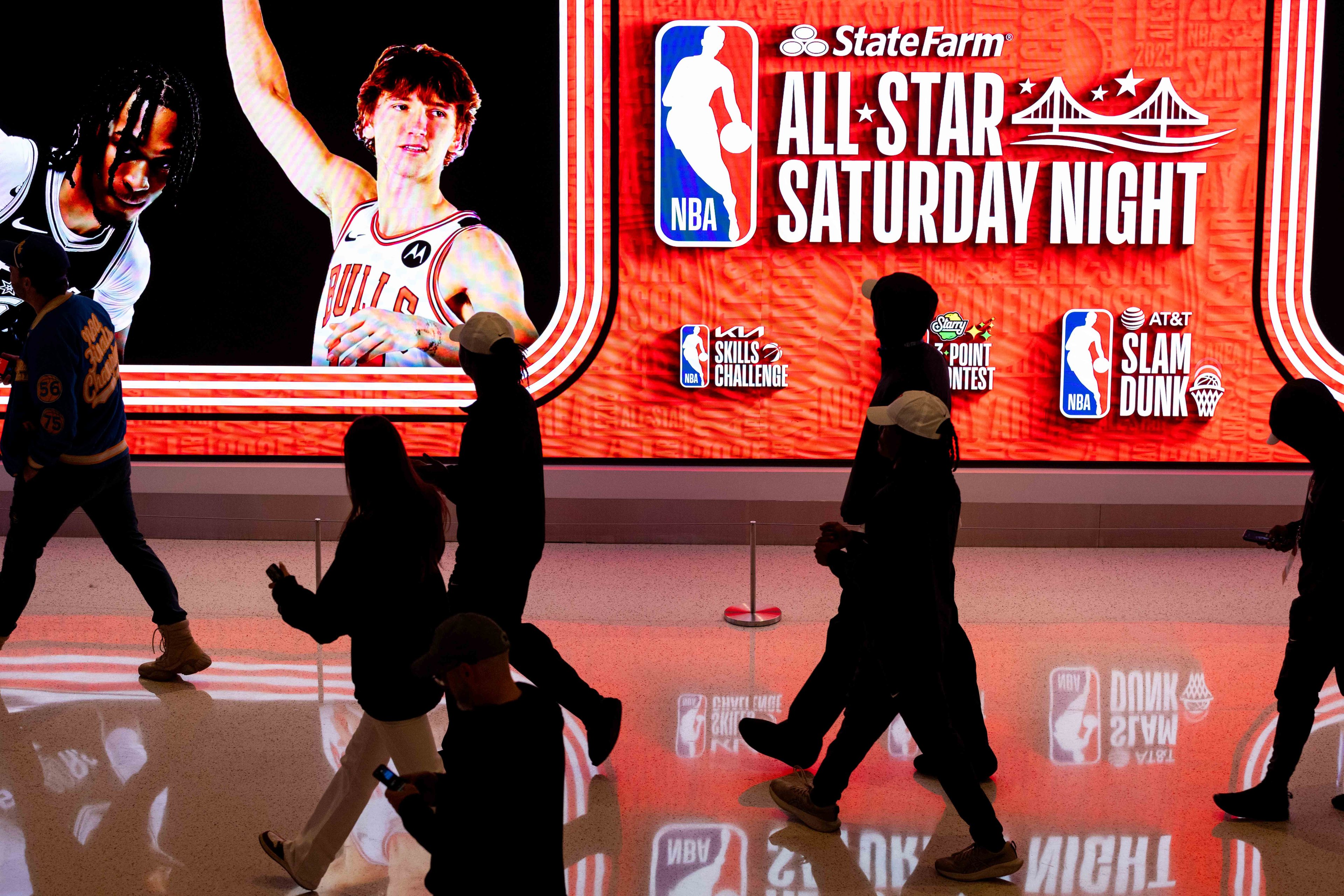 A group of people walks past a brightly lit sign featuring two basketball players, promoting NBA All-Star Saturday Night events like the Skills Challenge and Slam Dunk Contest.