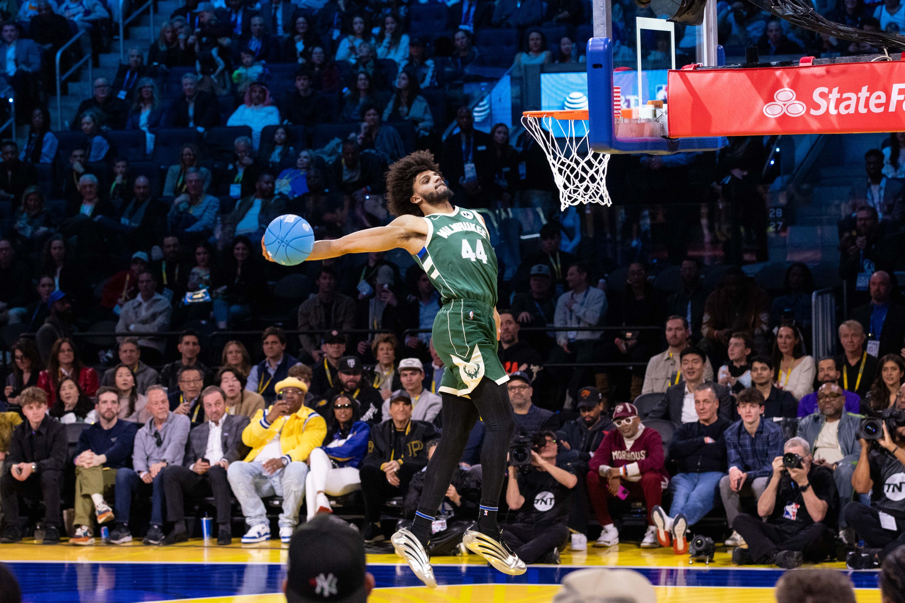 A basketball player in a green uniform performs an impressive slam dunk. The crowd watches intently as he soars towards the hoop, holding the ball high.