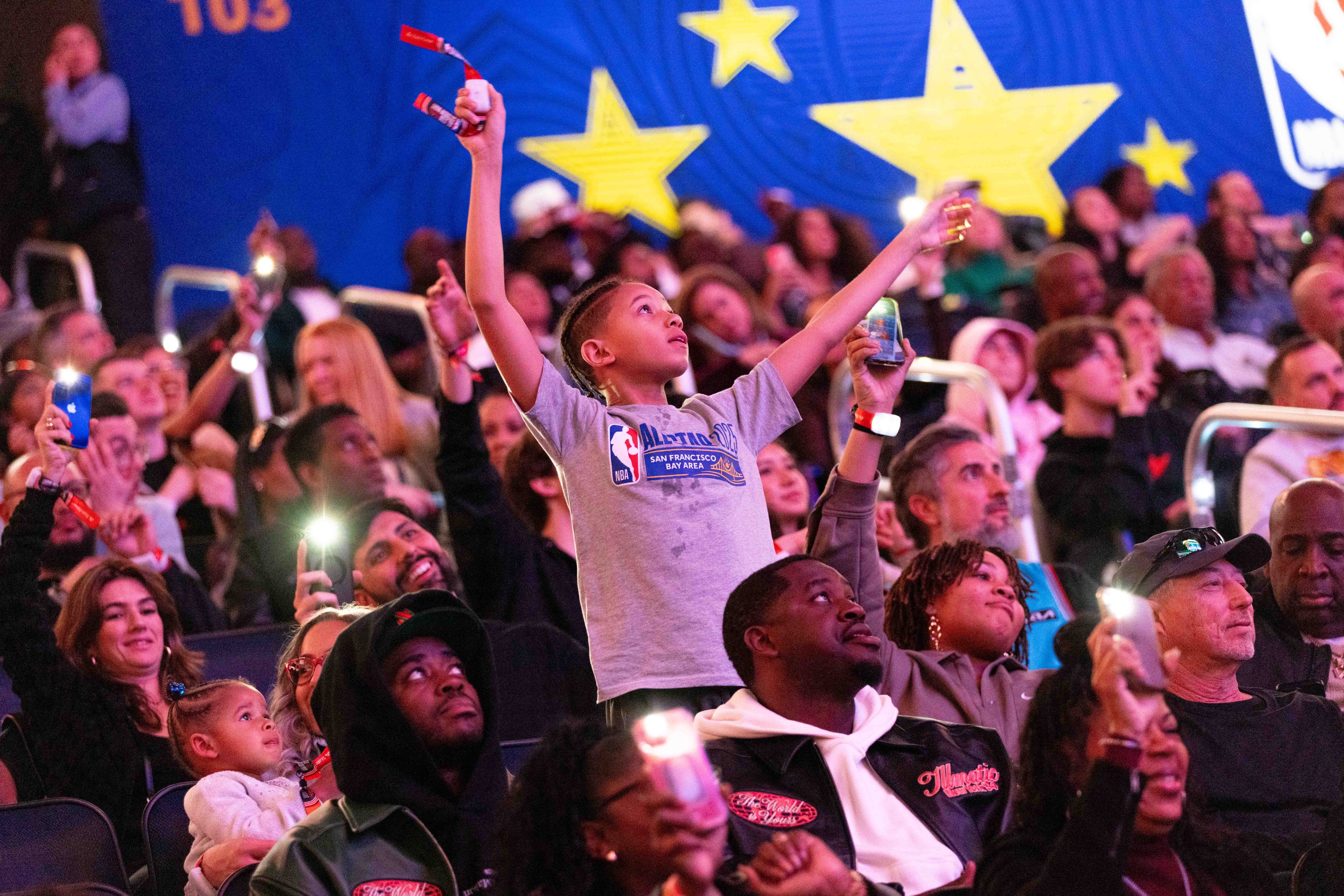 A crowd at an event is enjoying themselves, holding up lit-up phones and wearing bracelets. A child in the center has arms raised enthusiastically.