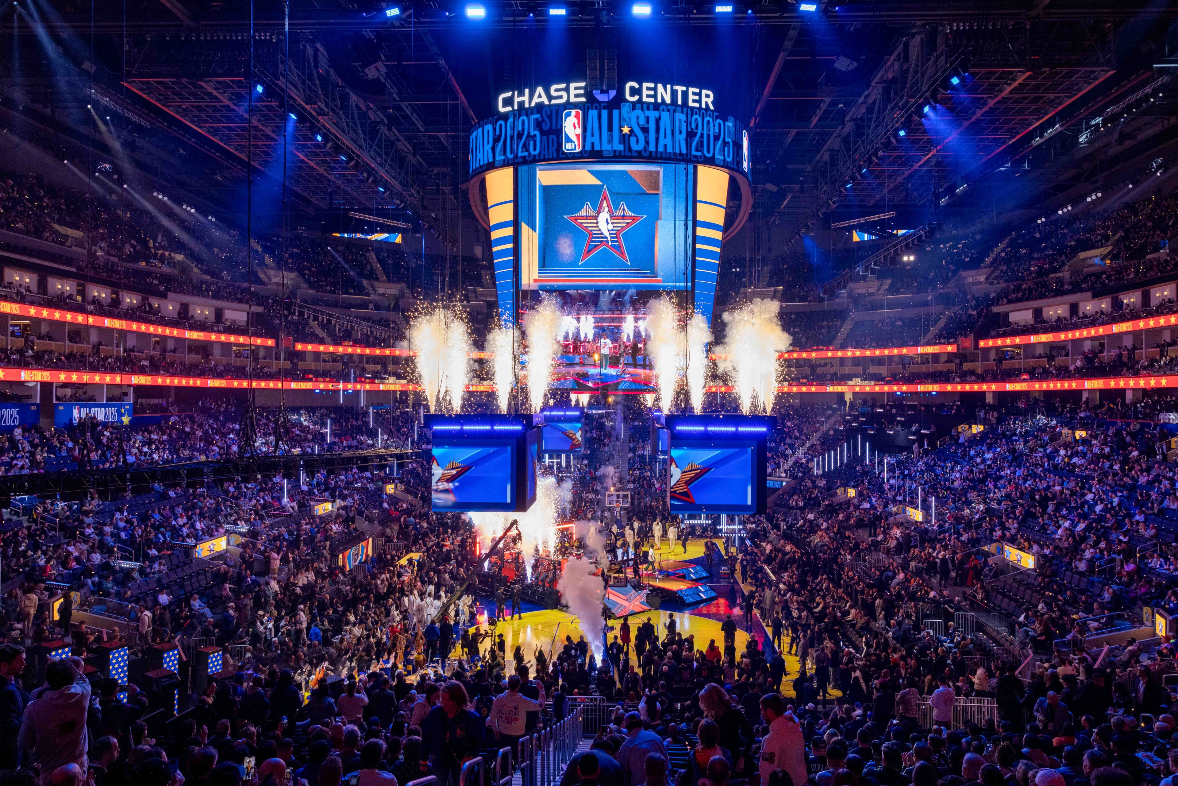 A packed basketball arena is vibrant with lights and fireworks, showcasing the NBA All-Star 2025 event. The crowd is energetic, and screens display logos.