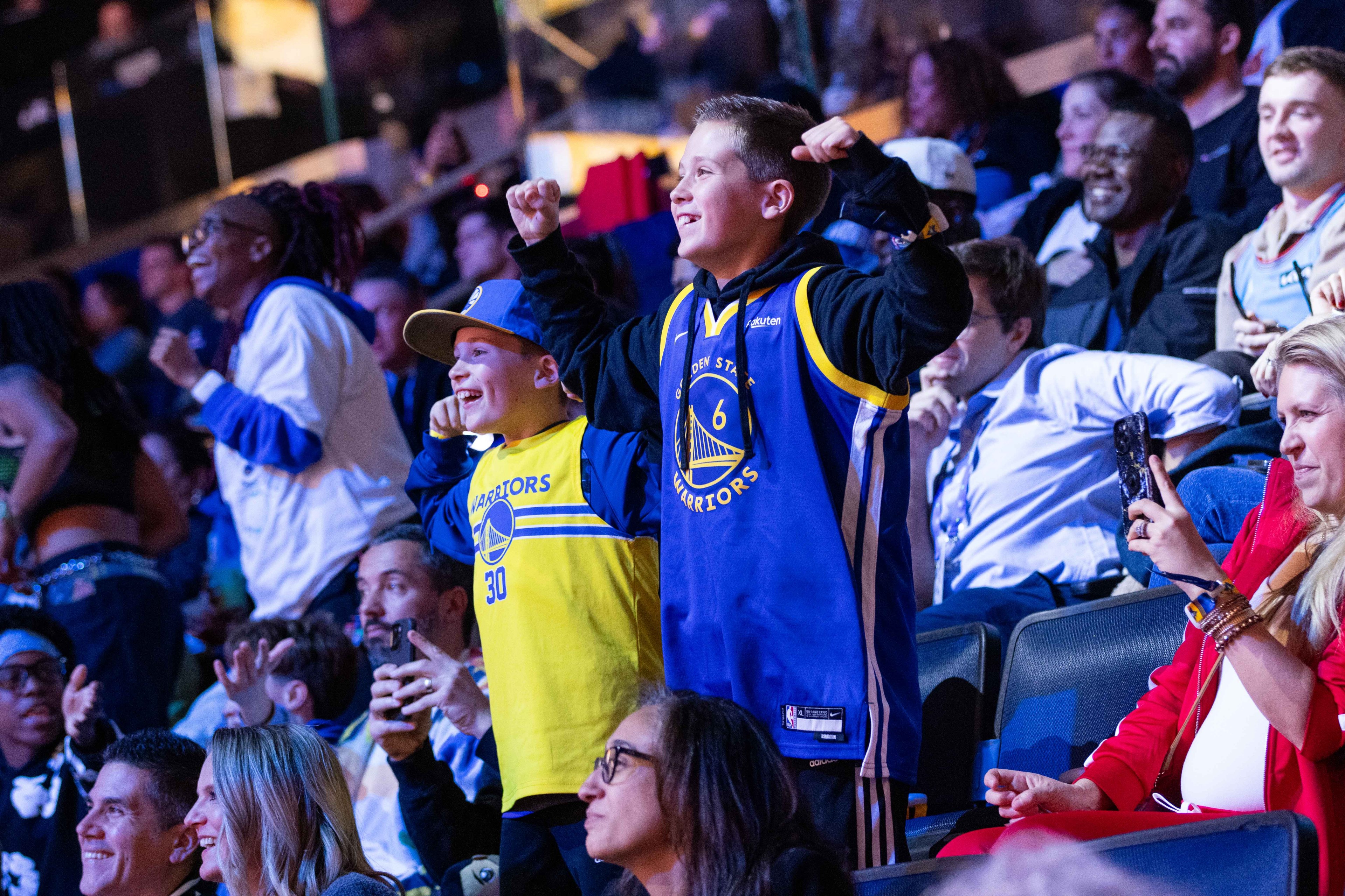 Two children wearing Golden State Warriors jerseys cheer excitedly in a lively crowd at a sports event. A woman in red holds a phone, capturing the moment.