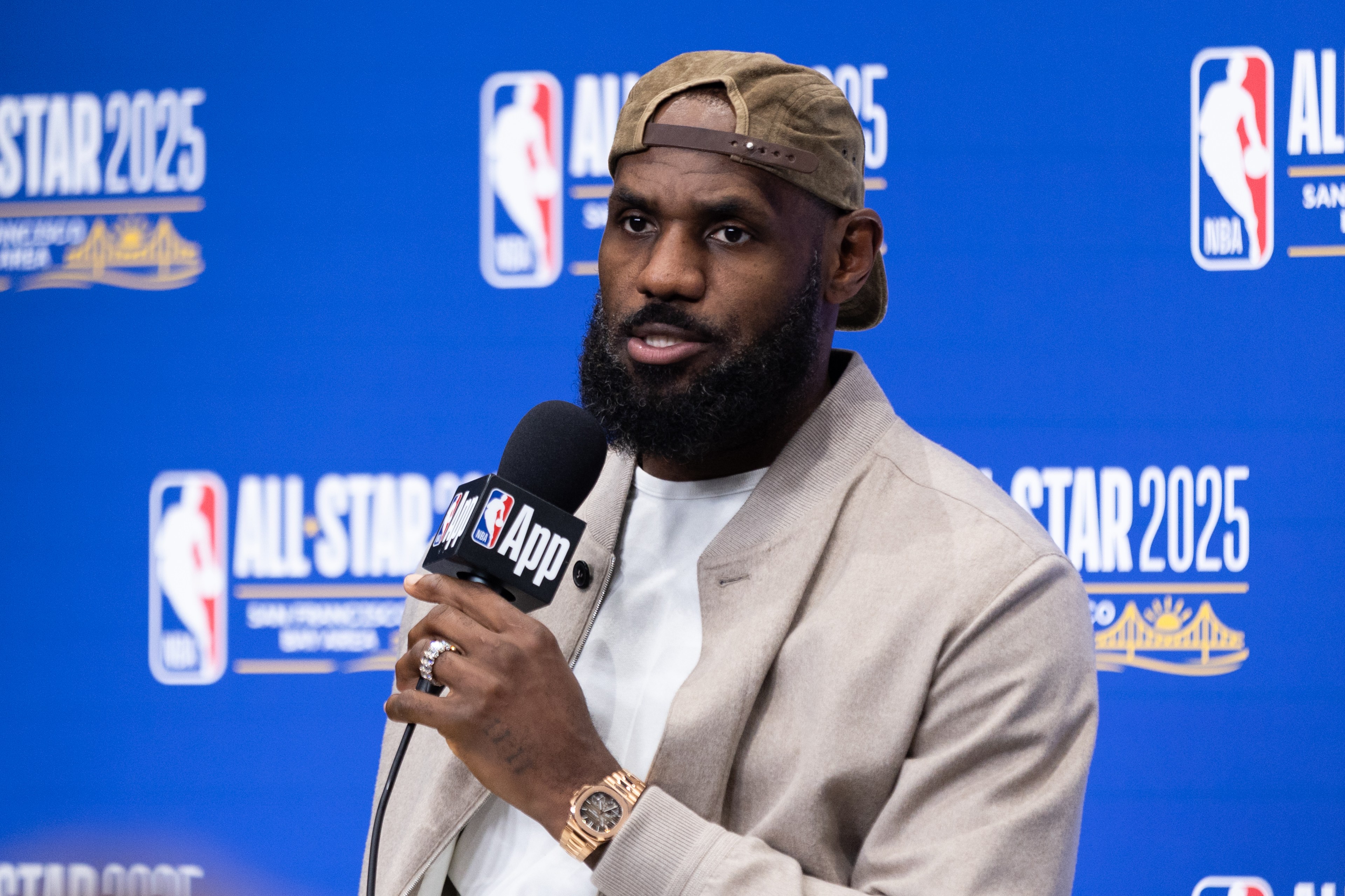 A man in a beige jacket and cap speaks into an NBA App microphone. The blue backdrop behind him displays &quot;All-Star 2025&quot; and related logos.