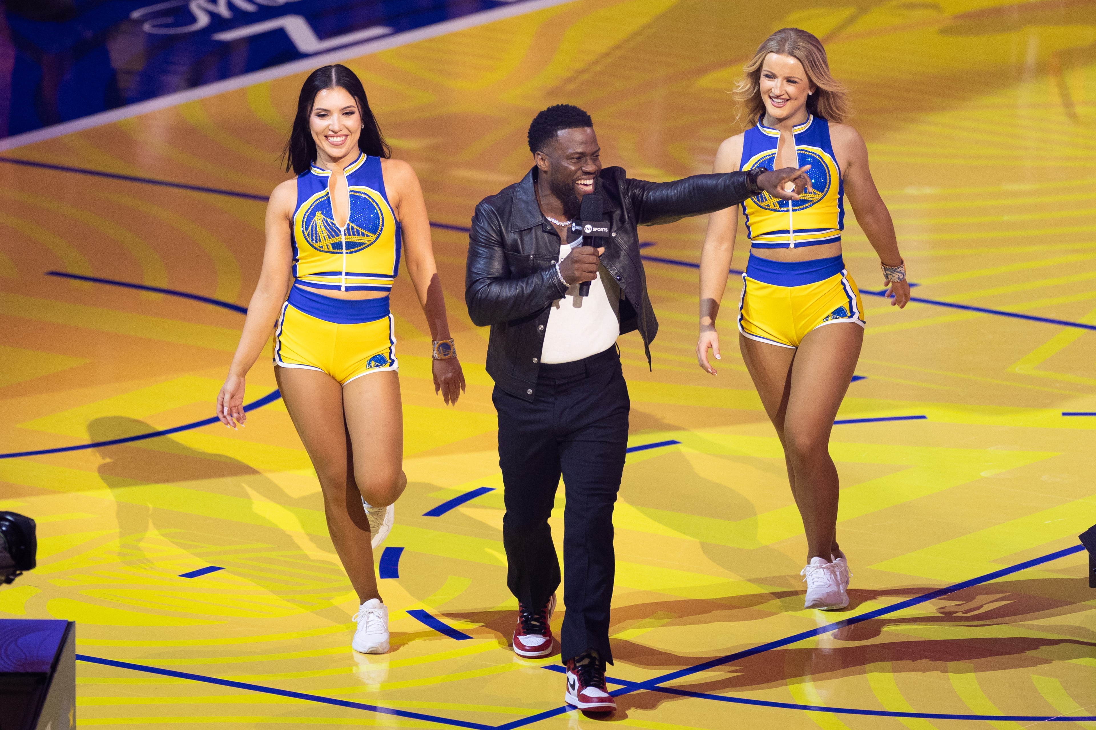 A person in a black jacket and sneakers walks on a sports court, holding a microphone, flanked by two cheerleaders in yellow and blue outfits.